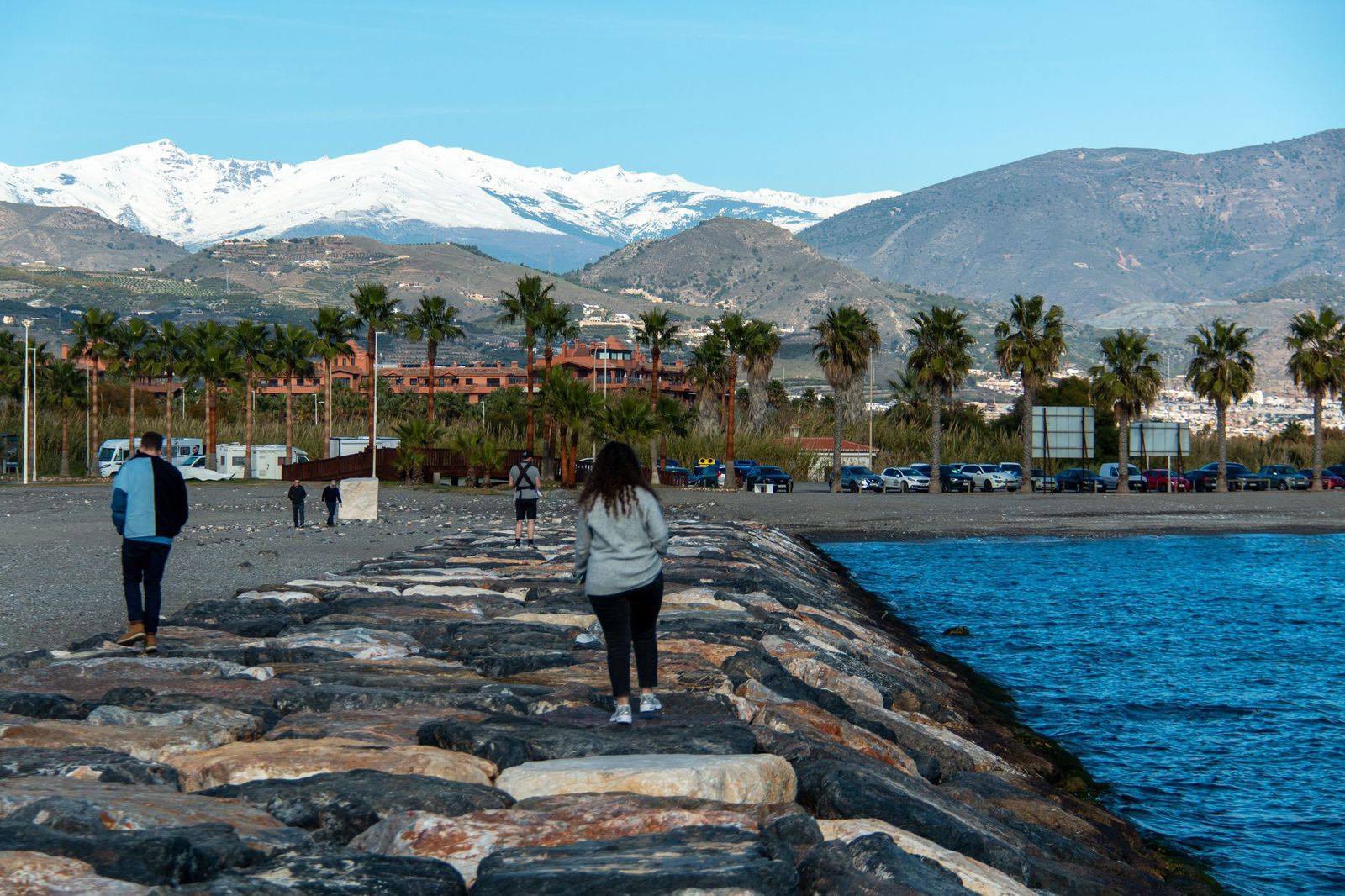 Varias personas pasean por el espigón de Playa Granada con Sierra Nevada de fondo