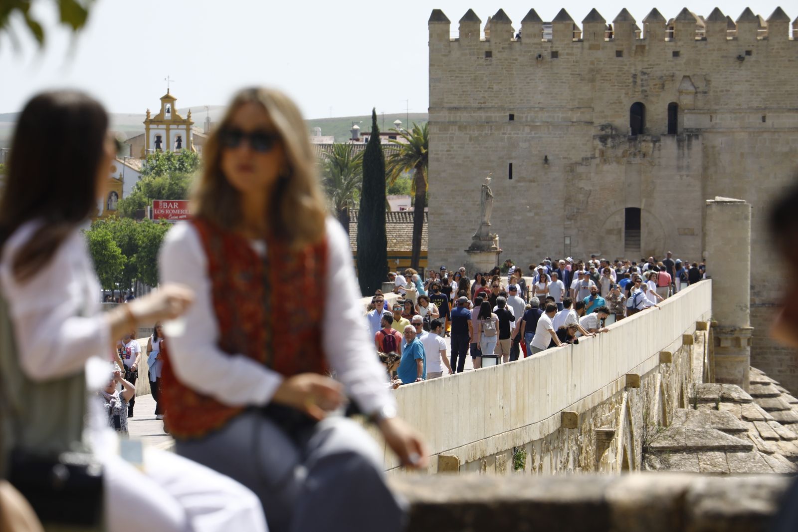 Dos chicas sentadas junto a un Puente Romano abarrotado de gente.
