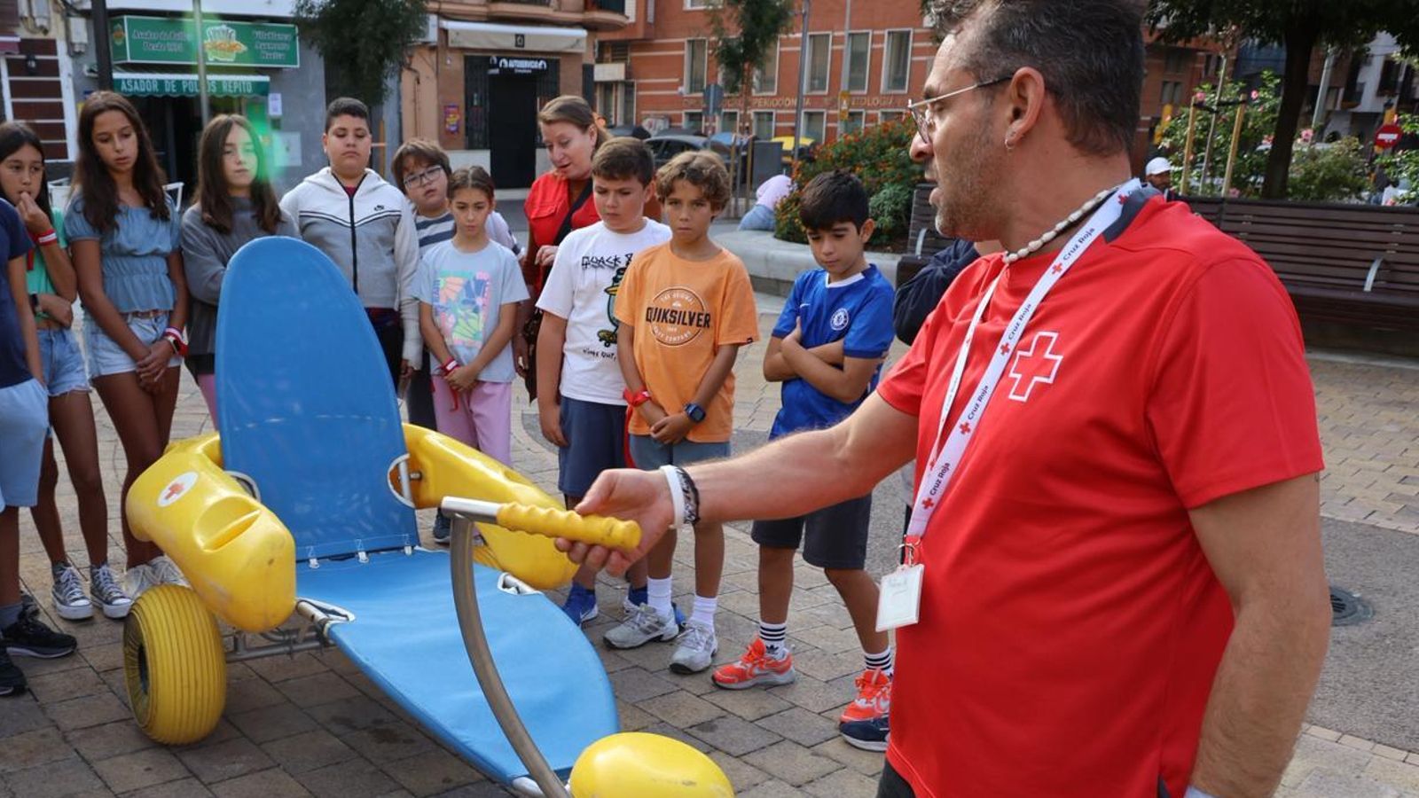 Un voluntario de Cruz Roja enseña a varios niños una de las sillas que se emplean en la playa para los baños inclusivos.