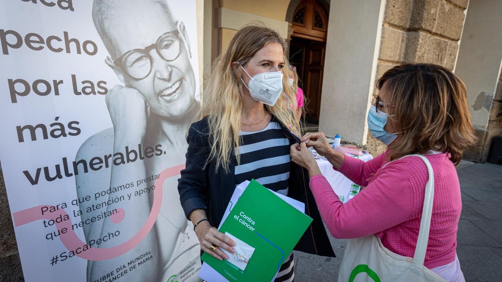 La vicepresidenta de la AECC en Cádiz pone un lazo en la solapa a la delegada del Gobierno andaluz en Cádiz en la plaza de San Juan de Dios.