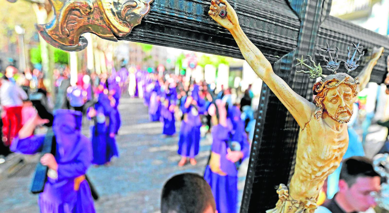 Primer plano de la cruz conventual de los Padres Capuchinos y, tras ella, nazarenos penitentes de la Hermandad de la Defensión.