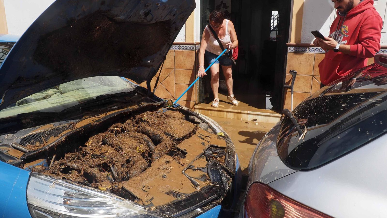 Un vecino de Lepe comprueba los daños en el motor de su vehículo, mientras otra achica agua del interior de una vivienda