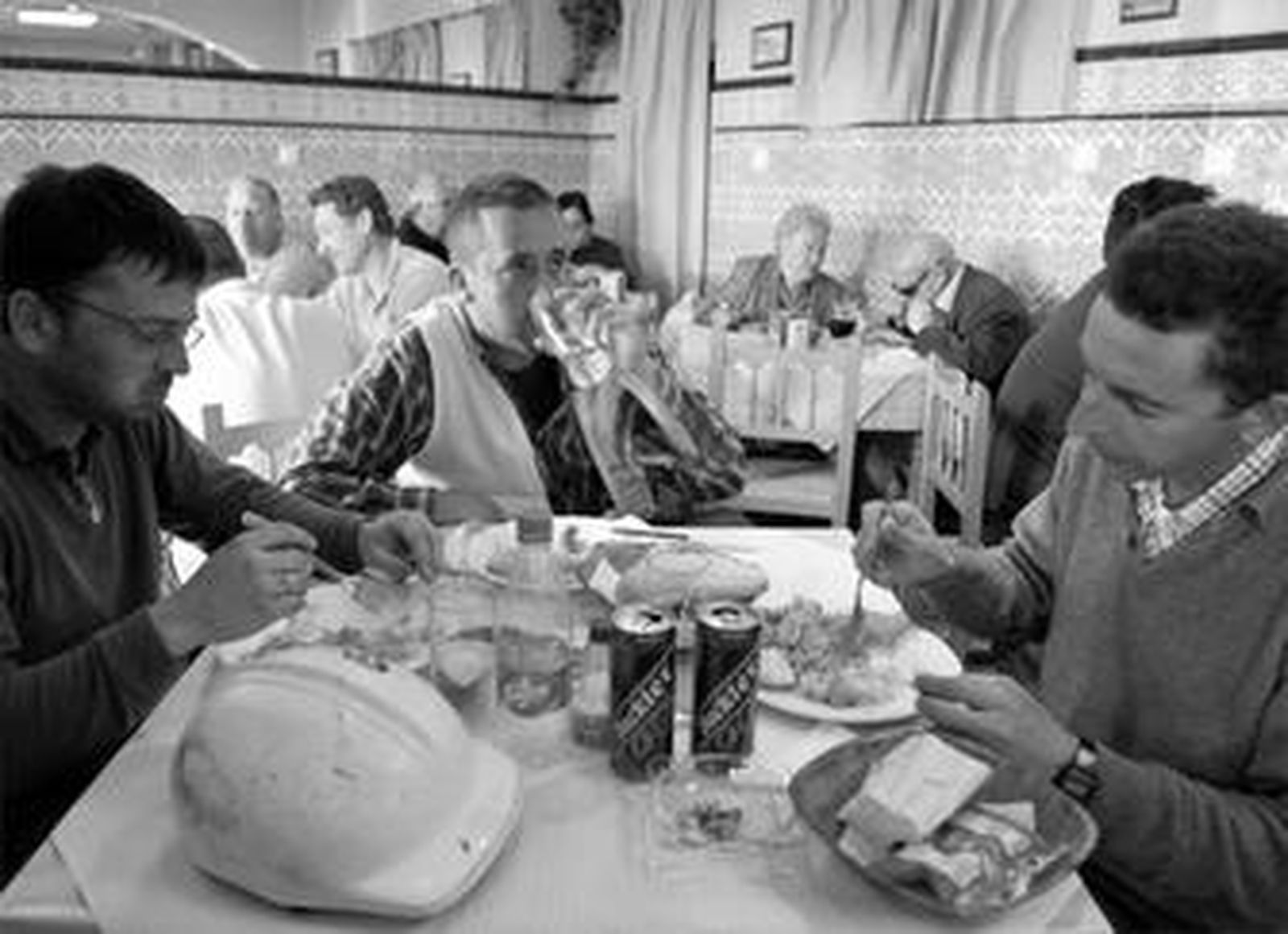 Tres trabajadores de la obra, comiendo en el restaurante Sancho Panza, con el casco encima de la mesa. Tras ellos, más.