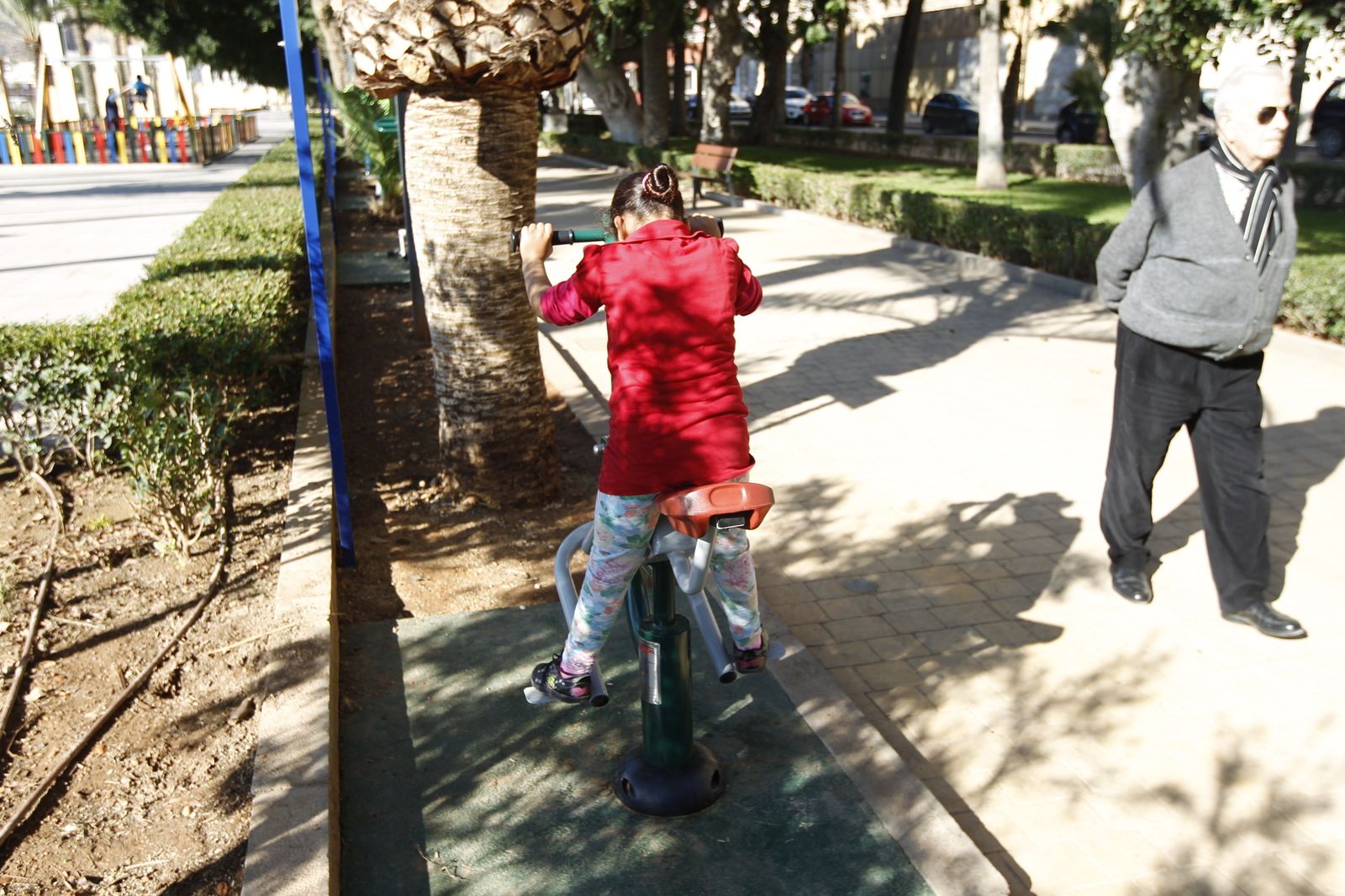 Un jubilado pasea delante de uno de los aparatos del área biosaludable mientras una menor lo usa.