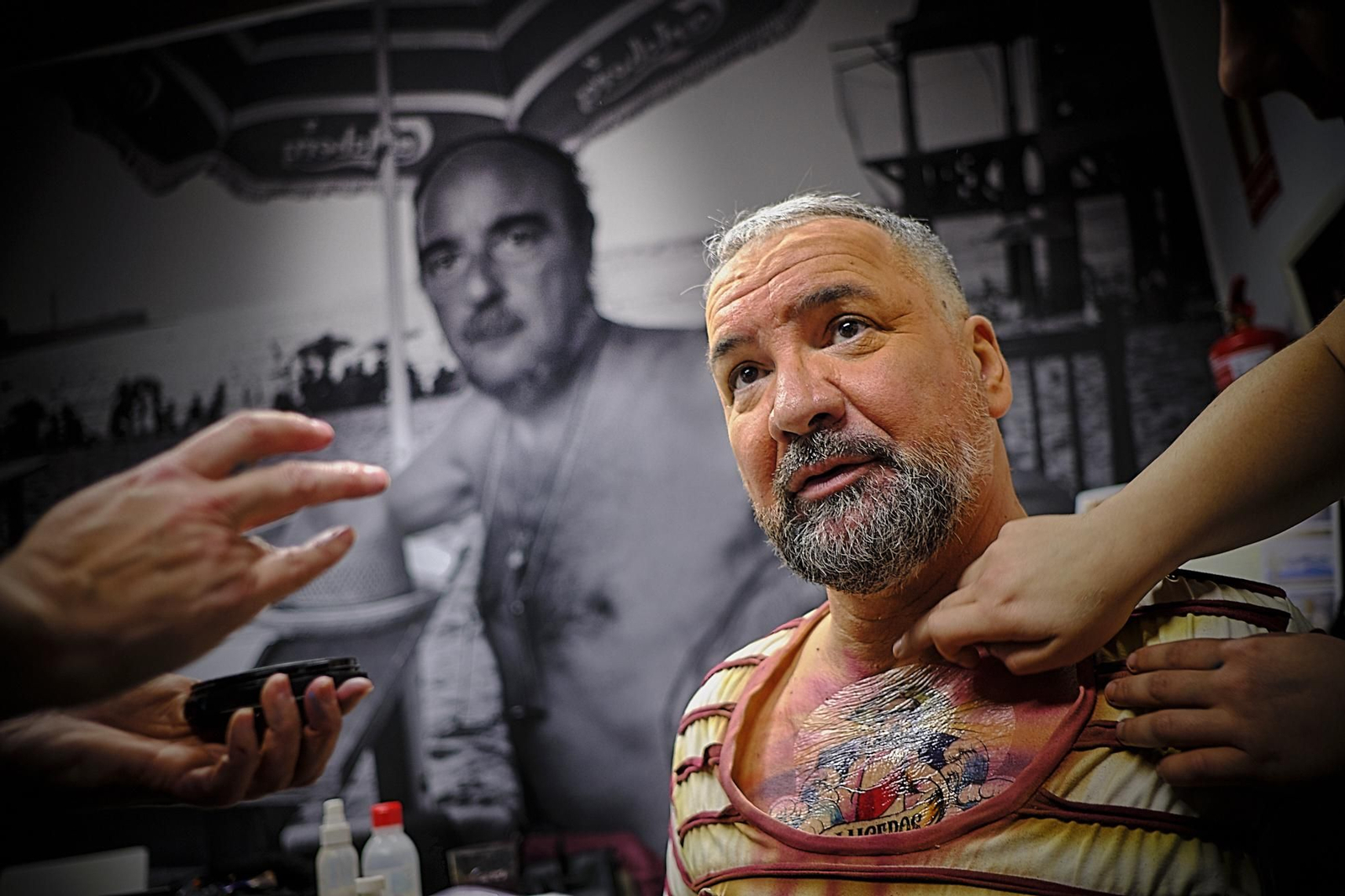 Ángel Subiela, director de la comparsa 'Los Luceros', recibe los últimos toques de maquillaje  junto a una imagen mural del escritor gaditano  Fernando Quiñones