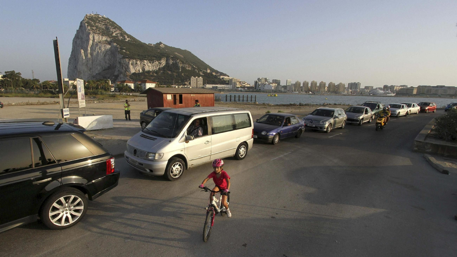 Una niña pasea en bicicleta junto a una cola de coches con el Peñón al fondo. Una niña pasea en bicicleta junto a una cola de coches con el Peñón al fondo.