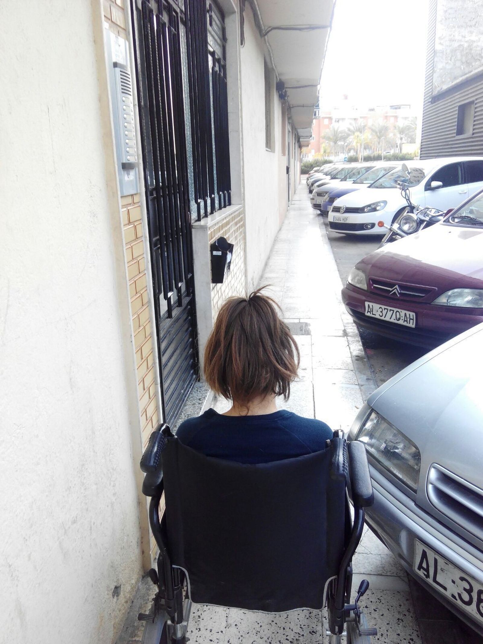 Una vecina en silla de ruedas entre dos coches estacionados muestra la escasa distancia que le dejan para pasar.