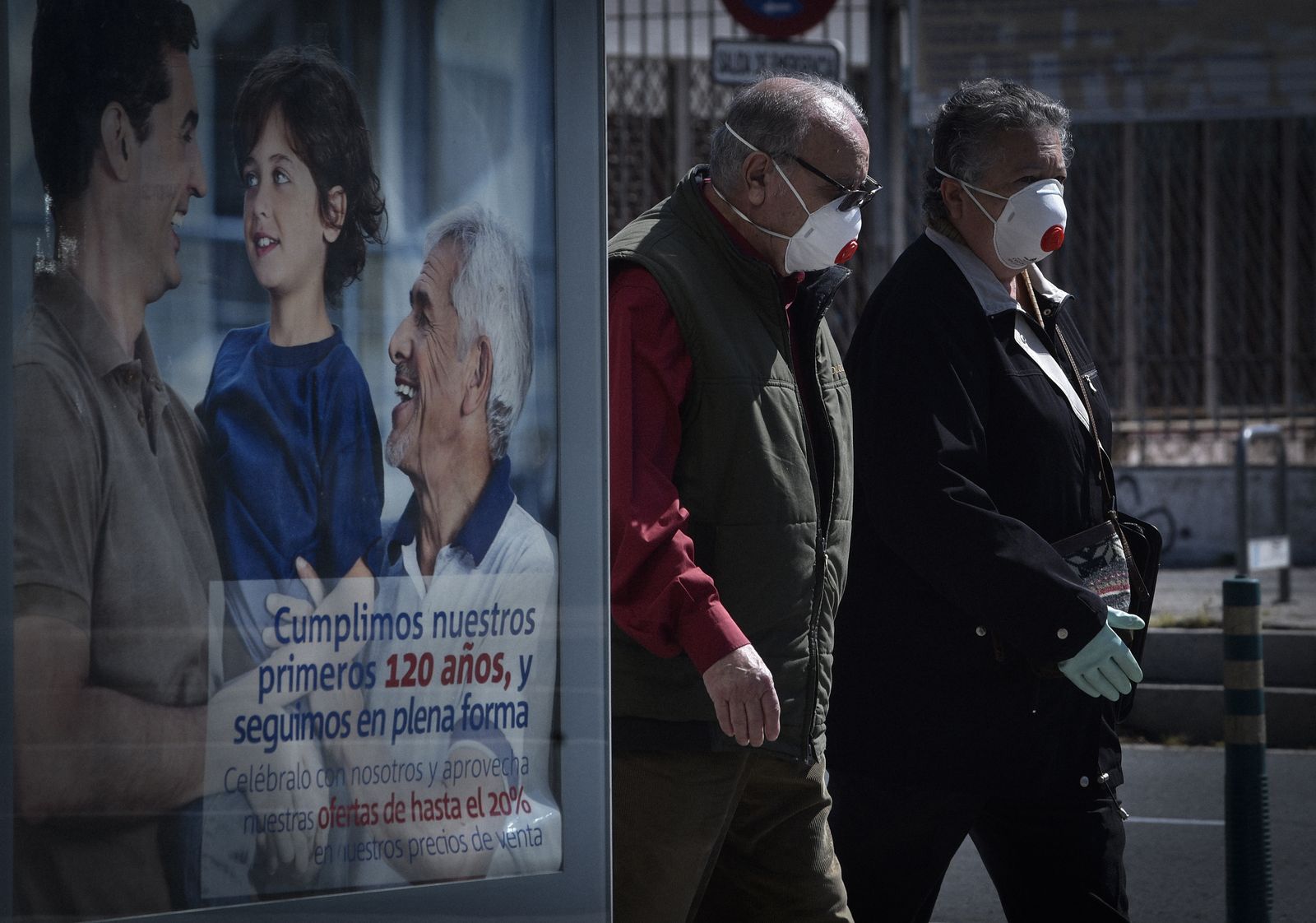 Dos personas mayores, con mascarillas de filtro y guantes, en una calle del barrio de la Macarena.