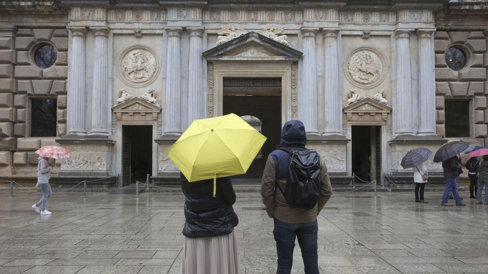 Un grupo de turistas observan bajo la lluvia la fachada del Palacio de Carlos V.