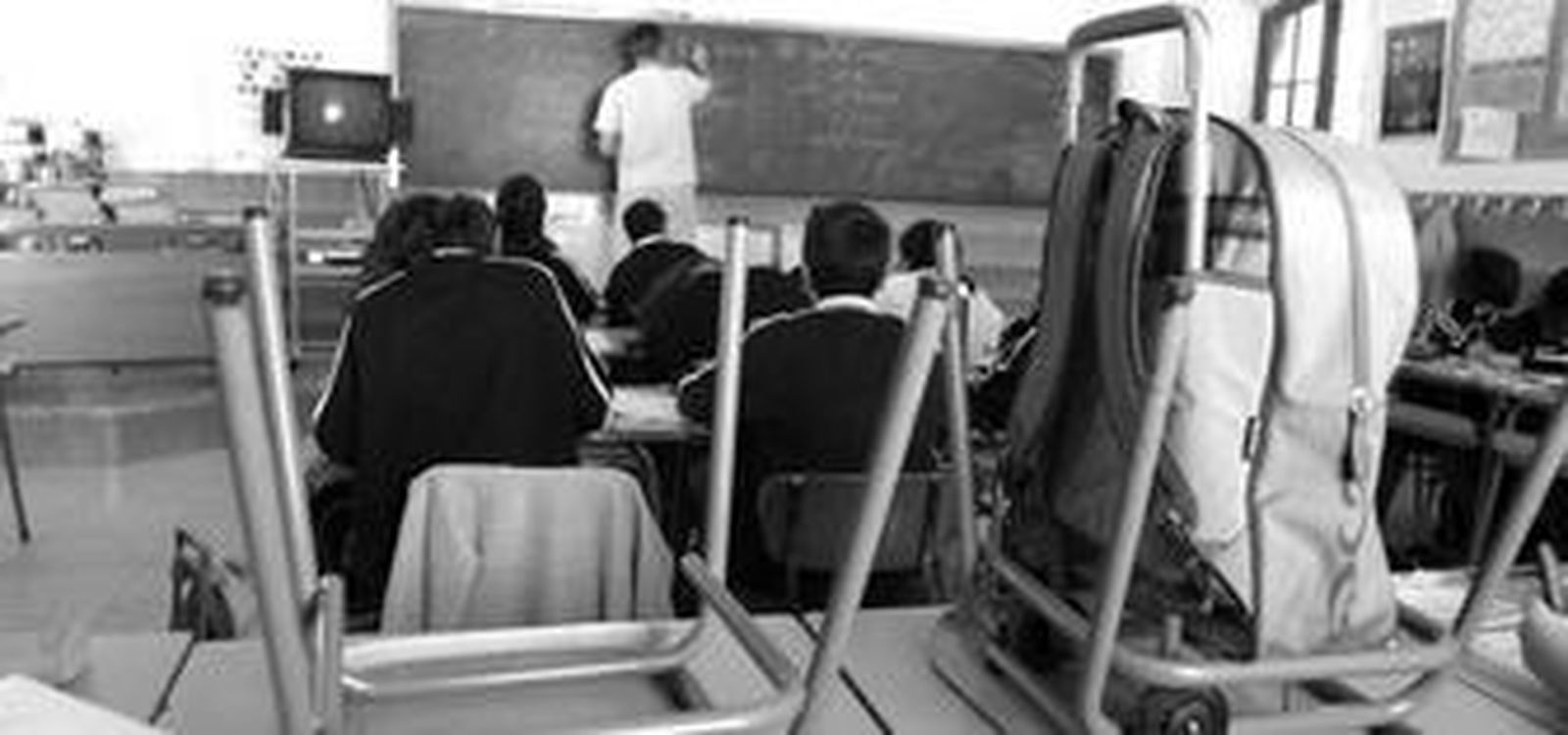 Pupitres vacíos en un centro, en el que un grupo de alumnos siguen las explicaciones del maestro.