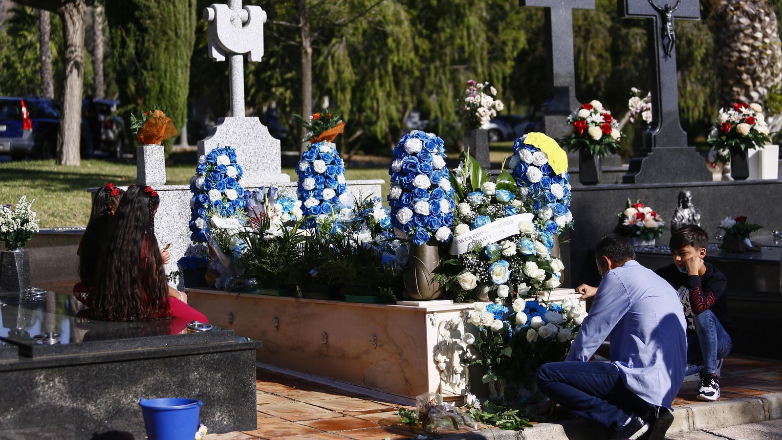 Un padre y sus nietos decora con flores azules y blancas la tumba de su hijo pequeño.