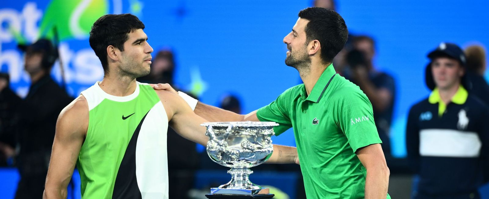 Carlos Alcaraz y Novak Djokovic se saludan antes de comenzar el partido con el trofeo delante.