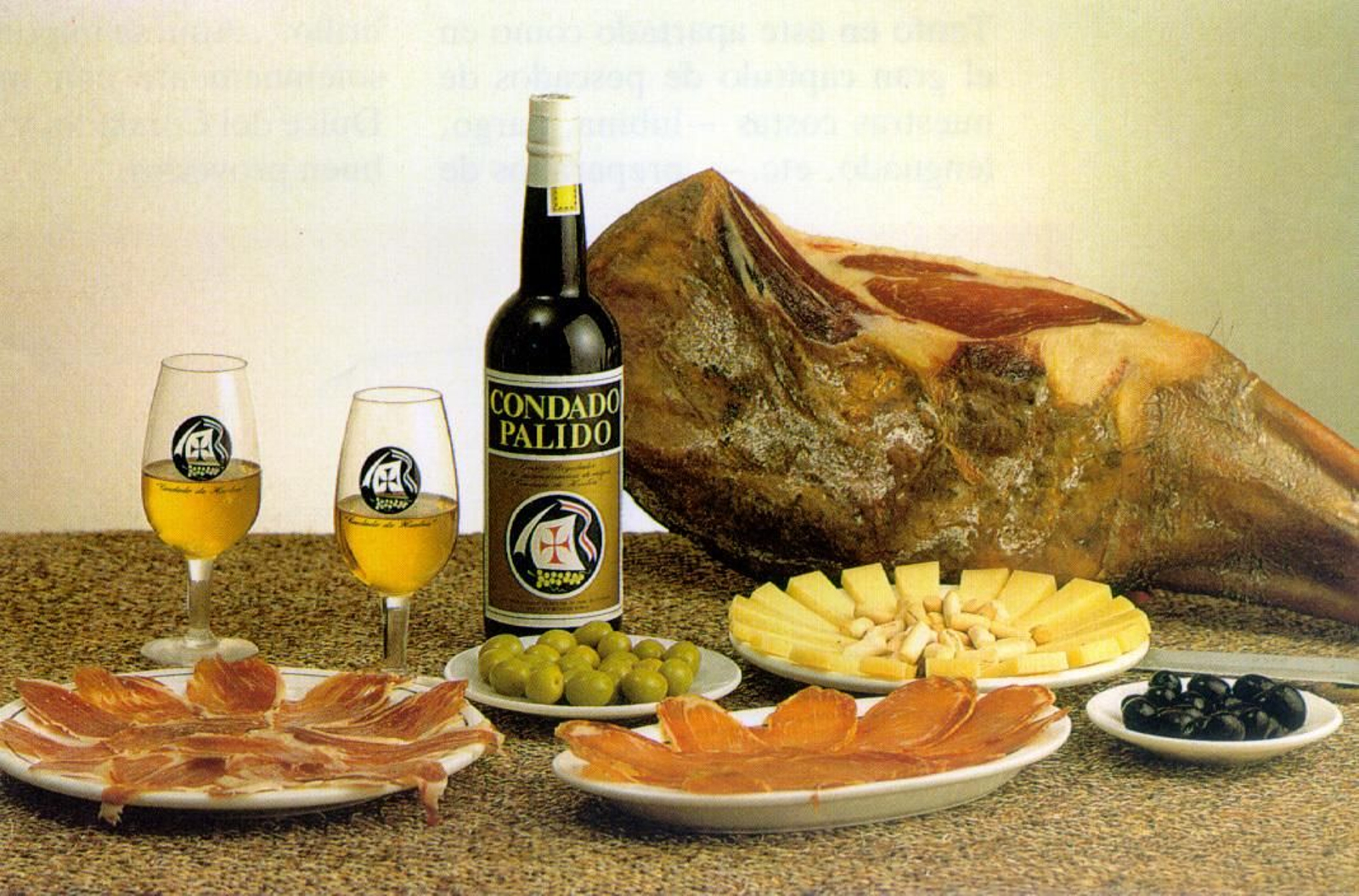 Una botella y dos copas de vino del Condado posan en una mesa junto a un jamón y platos de queso, lomo ibérico y aceitunas. Una botella y dos copas de vino del Condado posan en una mesa junto a un jamón y platos de queso, lomo ibérico y aceitunas.