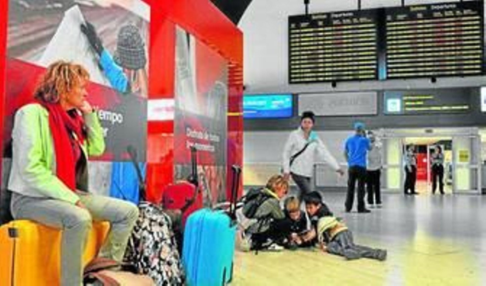 Una mujer espera sentada en su maleta mientras vigila a sus niños, que juegan a la videoconsola.