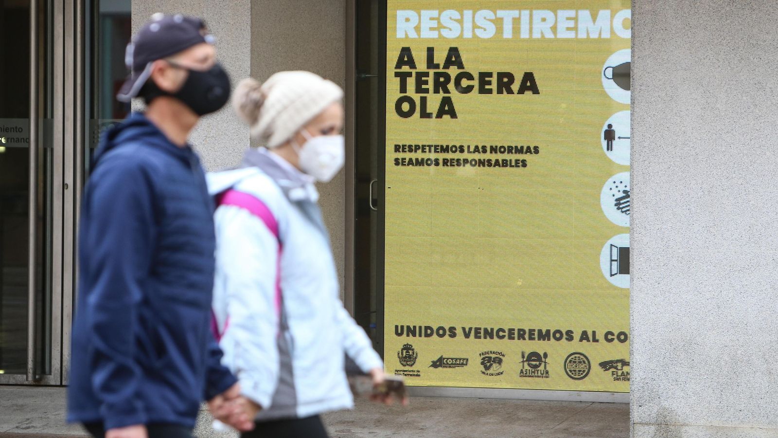 Una pareja con sus mascarillas pasa junto a un cartel del Ayuntamiento isleño alusivo a la pandemia.