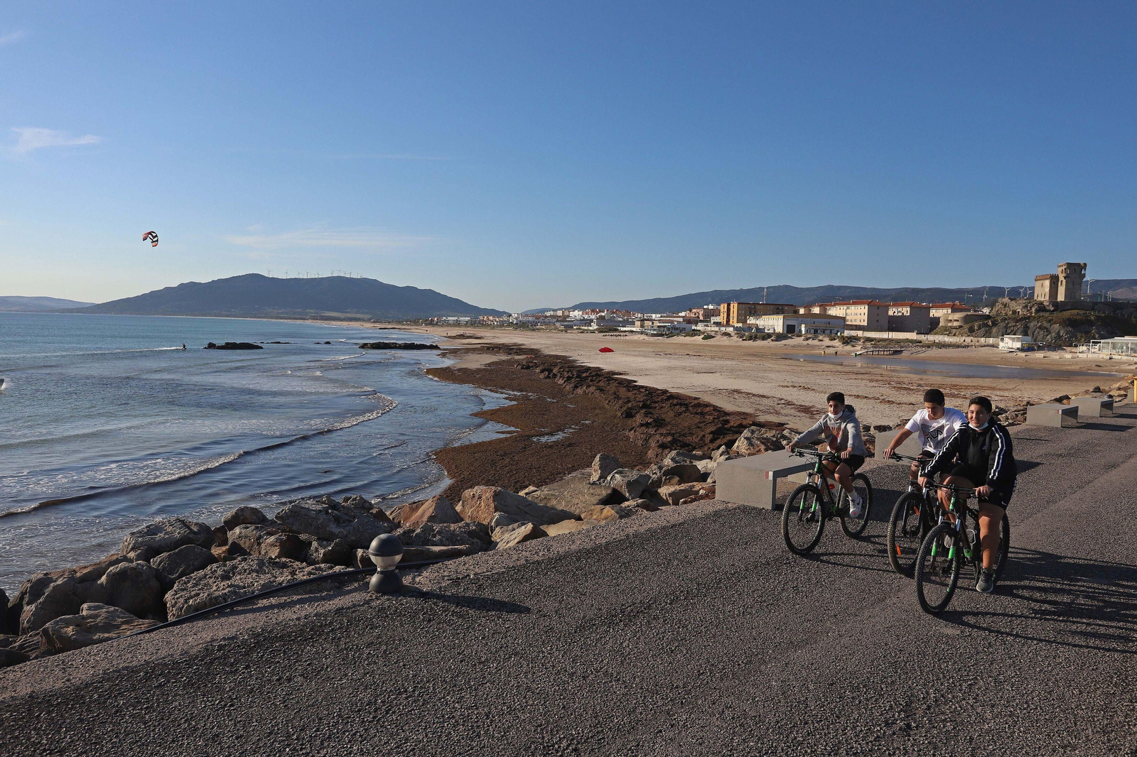 Tres jóvenes pasean en bici junto a la playa de Los Lances, en Tarifa, cubierta por el alga asiática.