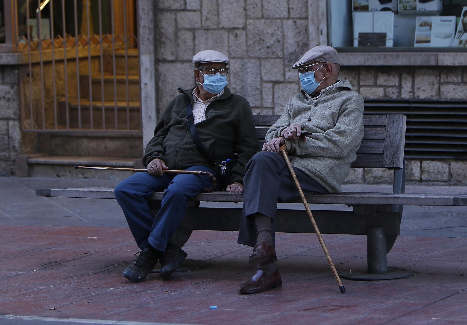 Dos ancianos conversan en un banco con mascarilla.