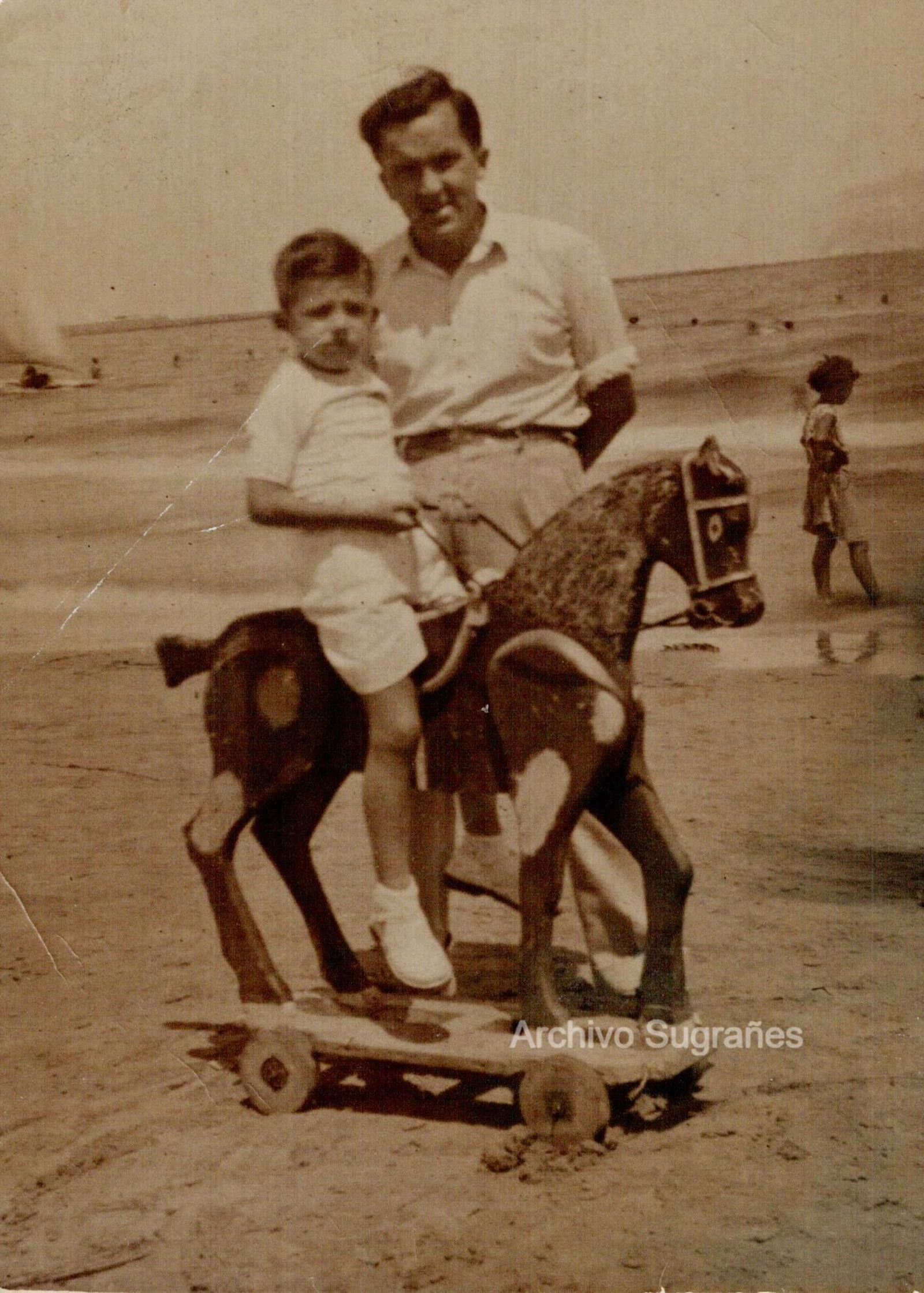 Un niño montado sobre un caballito de madera en la orilla de la playa junto a su padre.