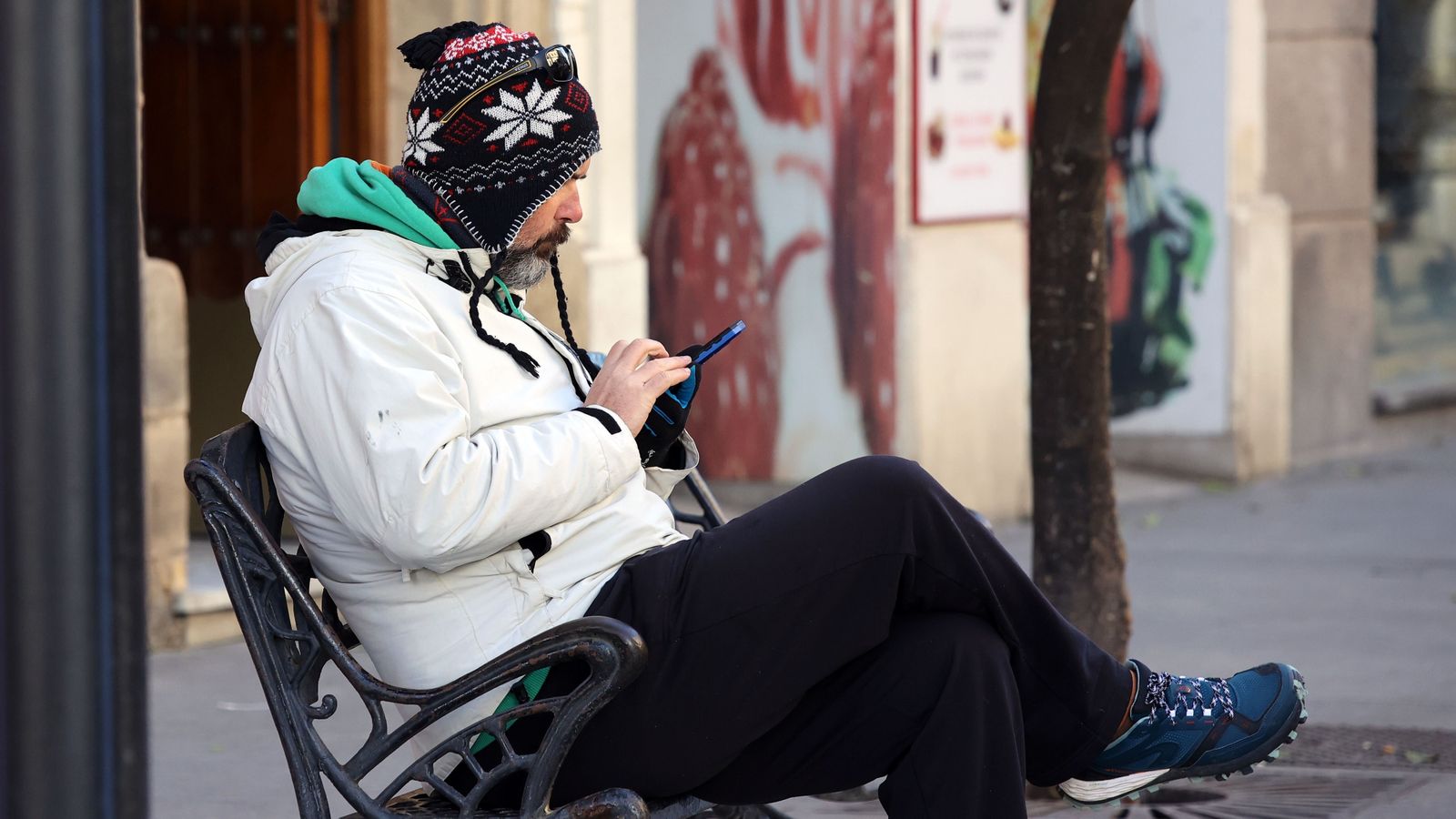 Una hombre abrigado con gorro consulta el móvil en un banco de la calle Larga
