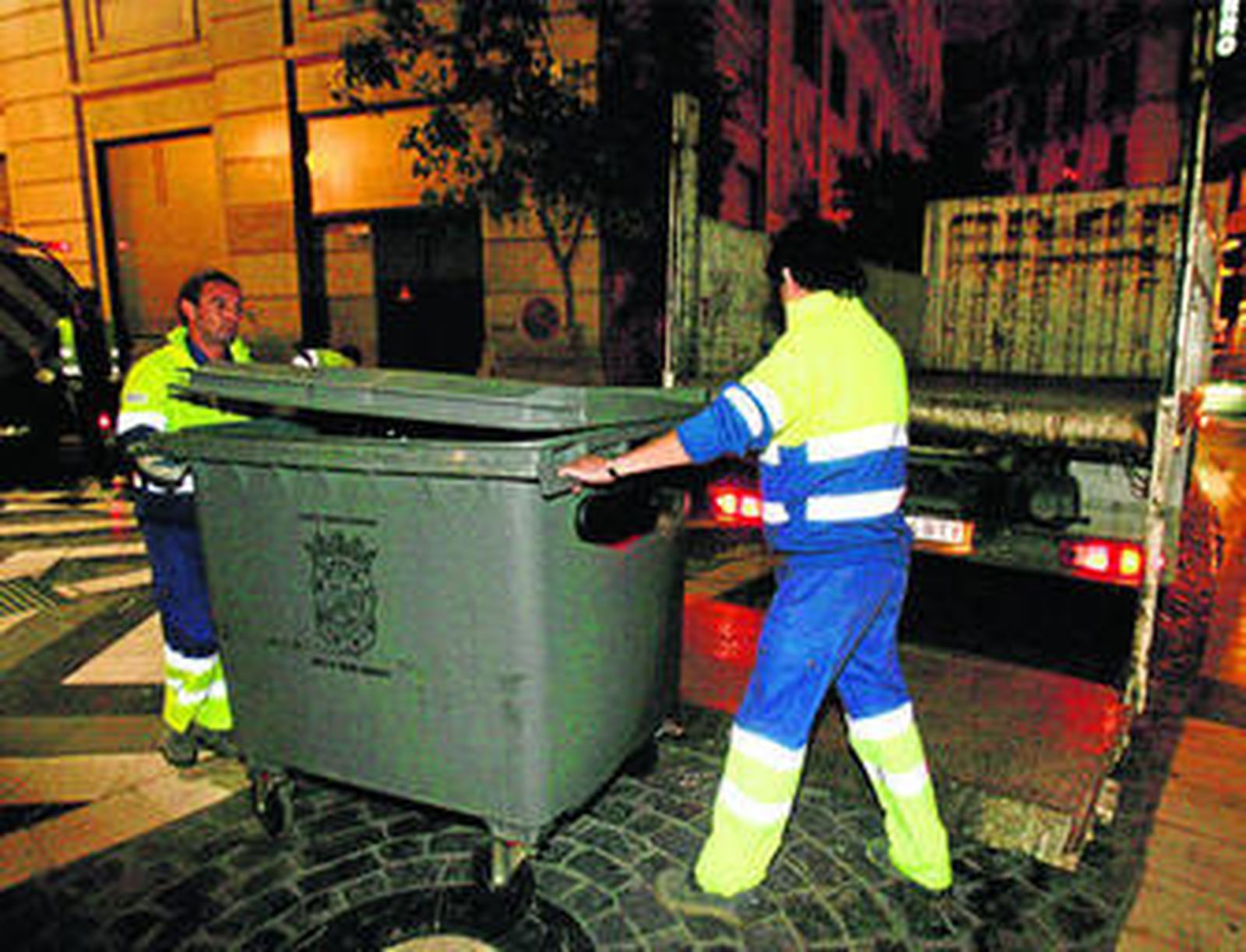 Dos operarios de Limasa recogen un contenedor de basura por la noche.