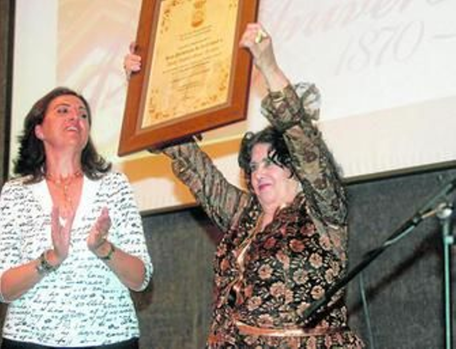 Bely Moya, junto a Rosario García, levanta emocionada el título que la acredita como Hija Predilecta.