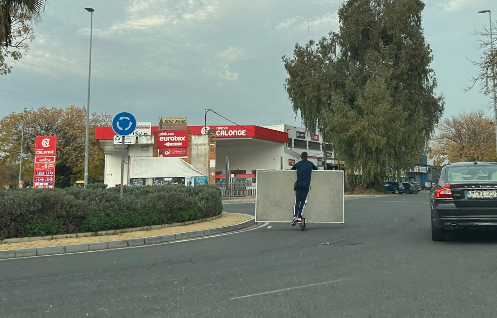 Un hombre lleva un panel cargado en un patinete pasando por una rotonda en Sevilla