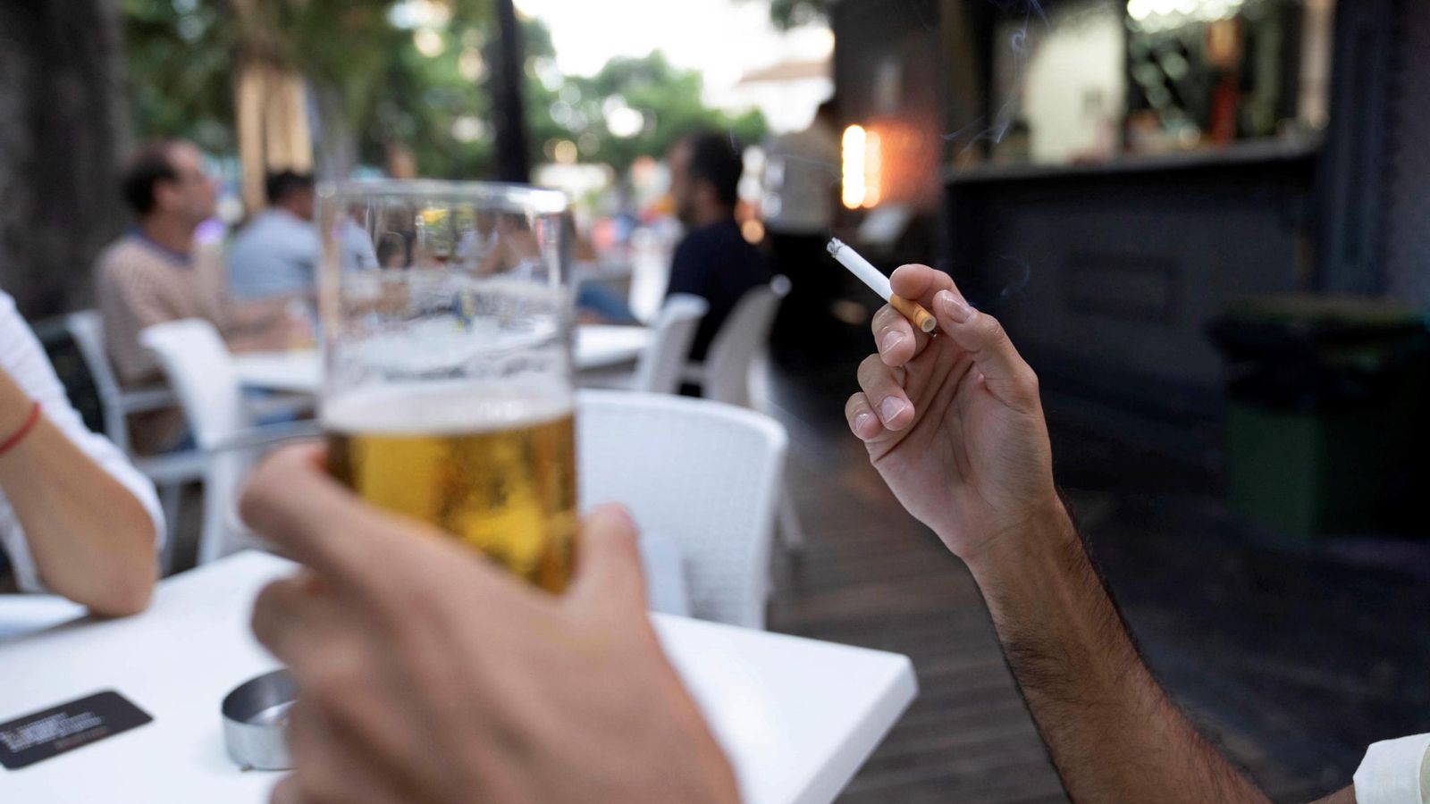 Un hombre sujeta un cigarro con la mano mientras en la otra sostiene una cerveza en la terraza de un bar.