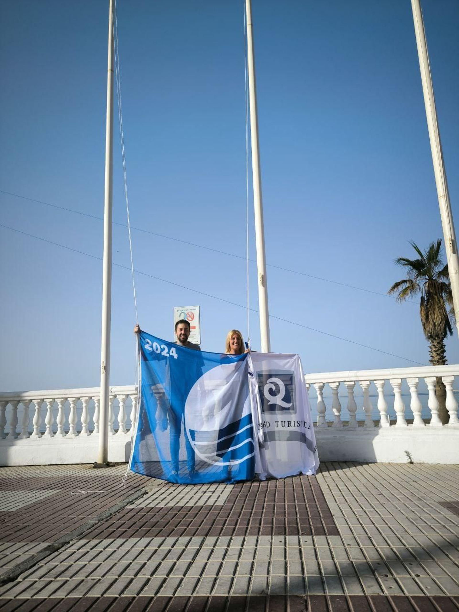 Ambos concejales sosteniento la Bandera Azul y el distintivo 'Q de Calidad Turística'