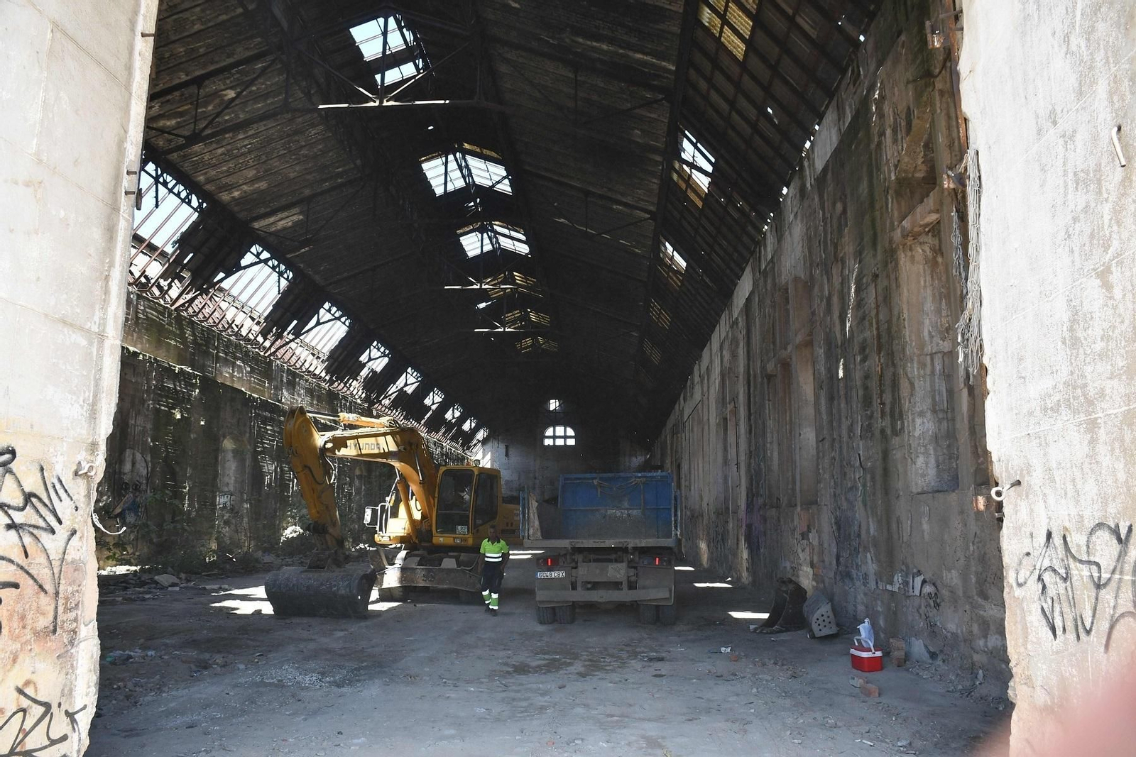 Un operario junto a una excavadora y un camión en el interior del edificio.