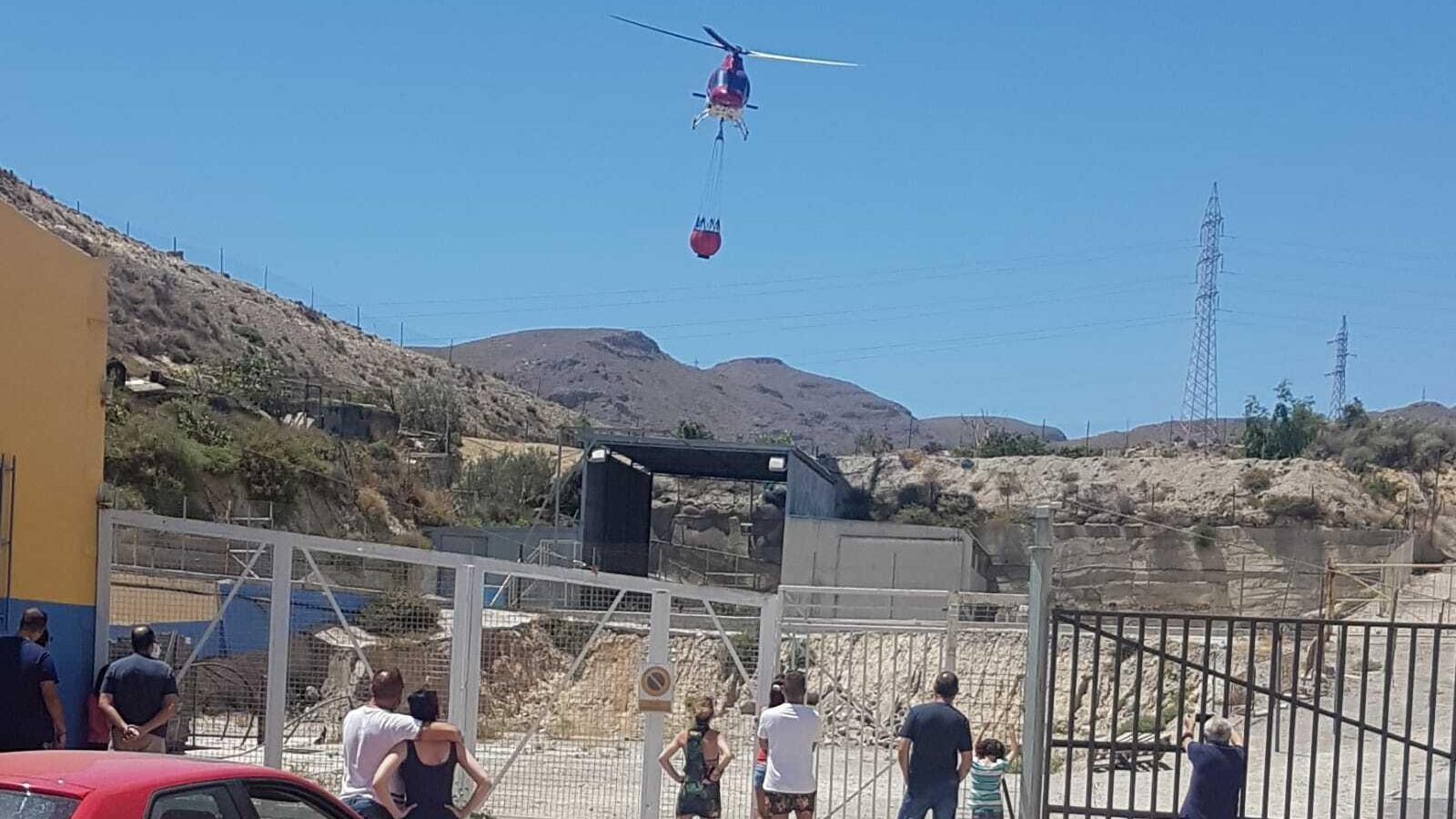 Un helicoptero de extinción recoge agua de un embalse en los aledaños al incendio, ante la atenta mirada de personas que circulan por la N-340a
