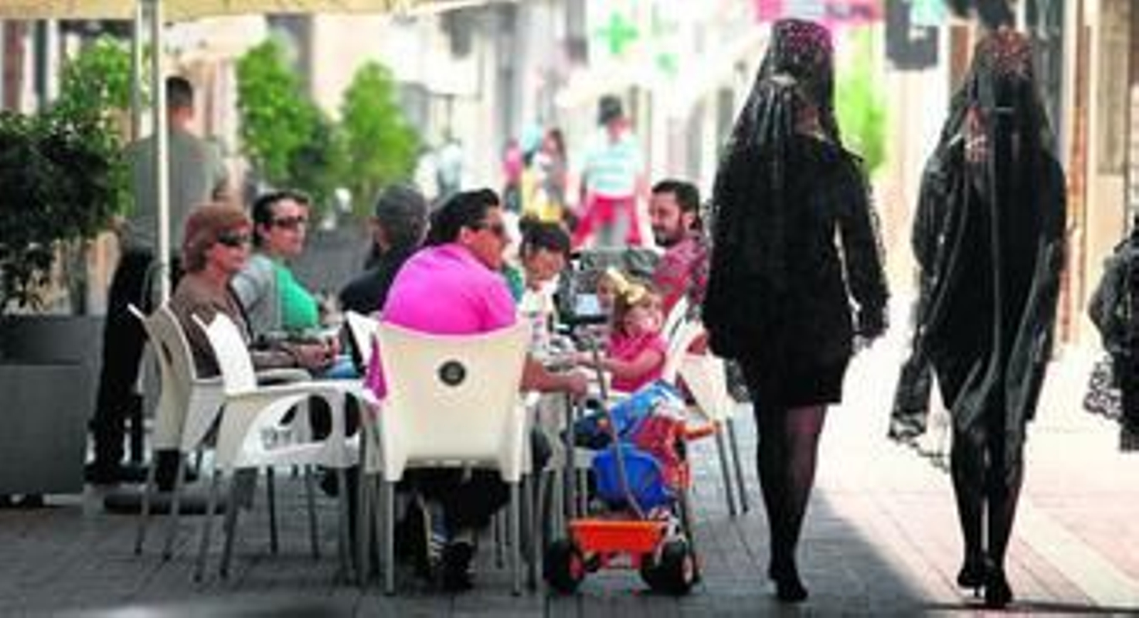 Jóvenes ataviadas con mantilla pasan junto a las terrazas donde se refrescan los ciudadanos en la calurosa mañana del Jueves Santo.