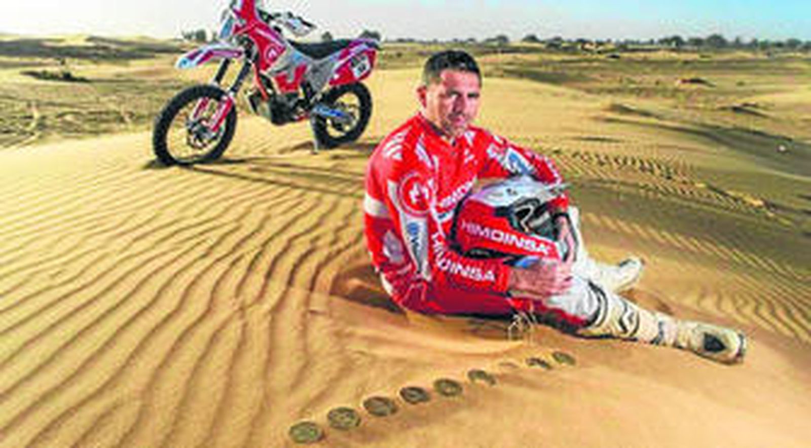 Miguel Puertas posa en la arena por delante de su moto y junto a las nueve medallas que acreditan las veces que acabó el Dakar.