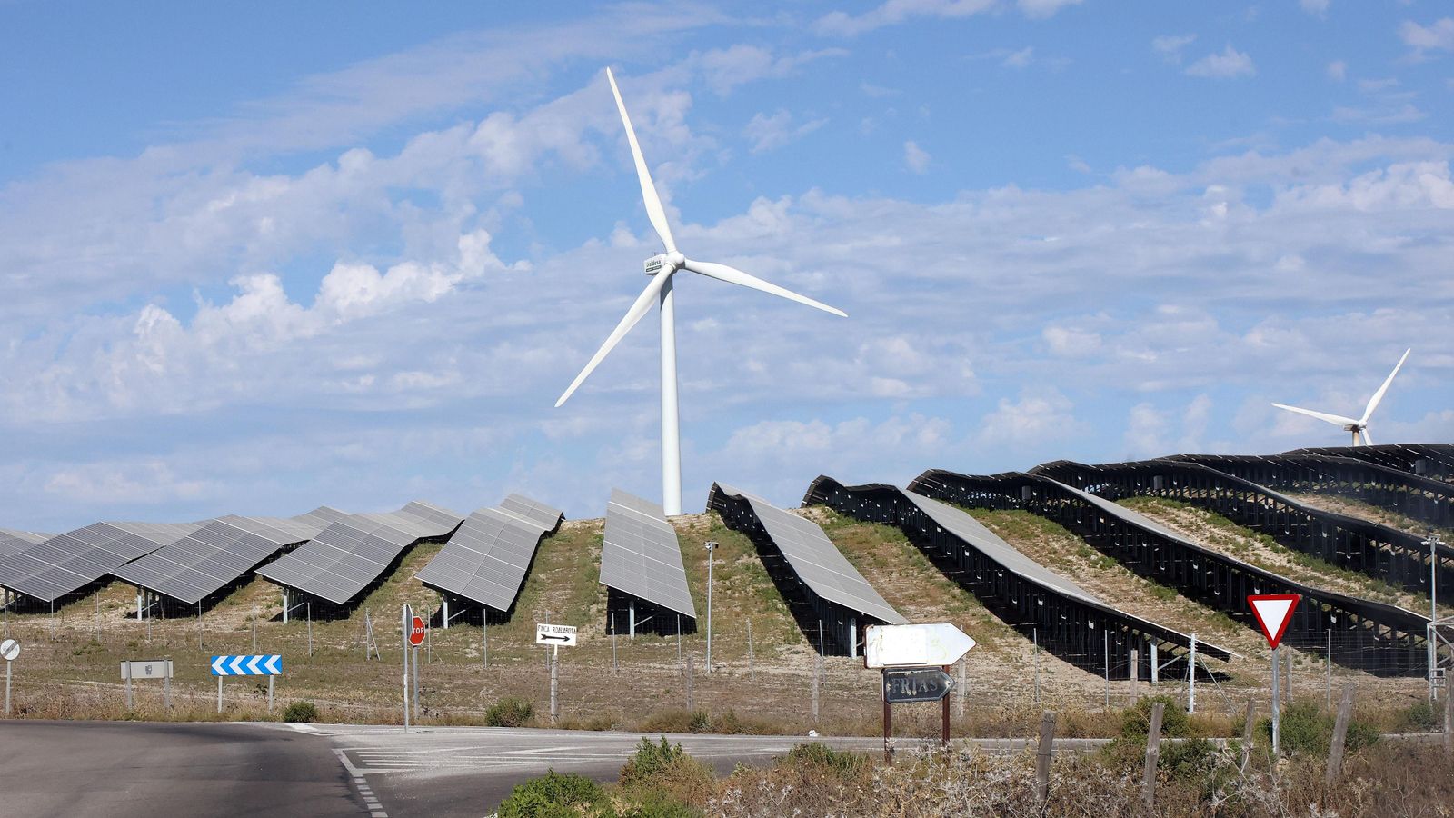 Una pequeña loma ocupada por paneles fotovoltaicos con aerogeneradores al fondo.
