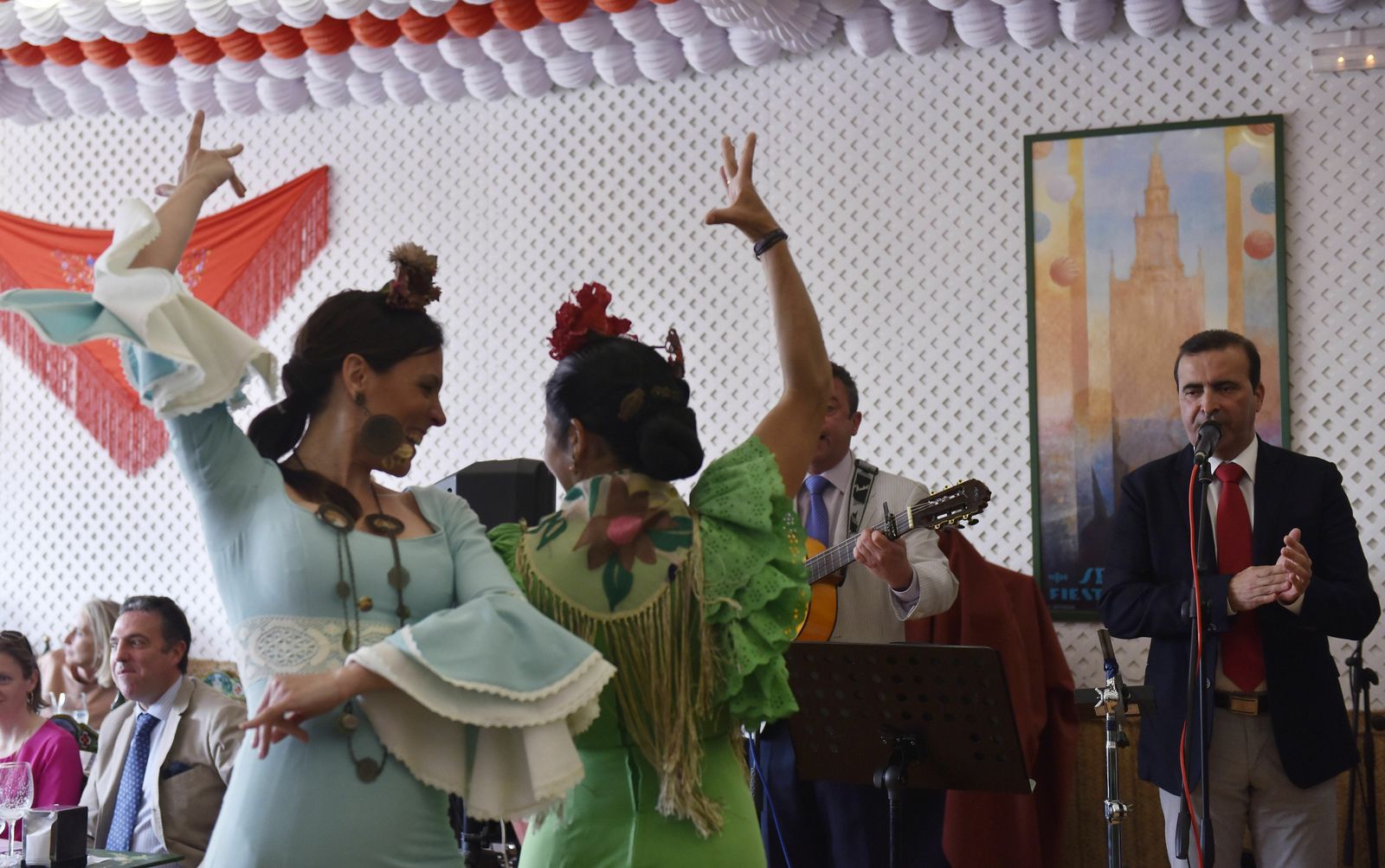 Dos jóvenes bailan sevillanas mientras un grupo ambienta la caseta con sevillanas.