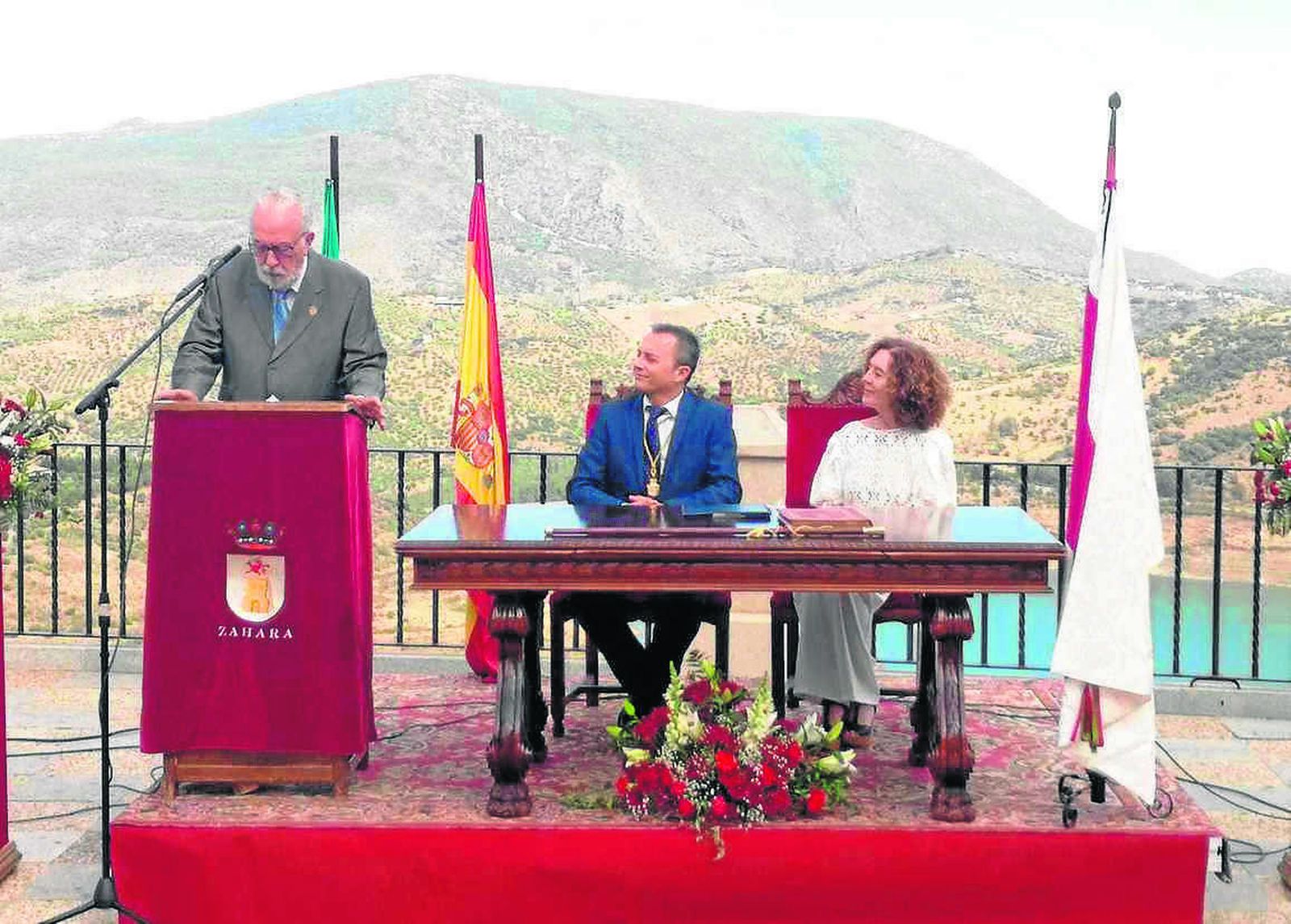 Rodríguez Becerra lee su discurso. Sentados, el alcalde de Zahara y la secretaria. Al fondo, la sierra de Lijar.