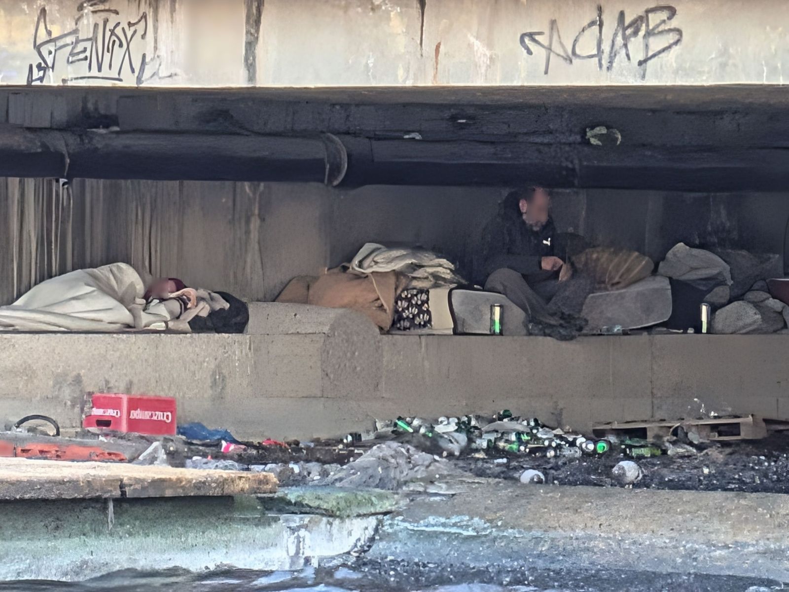 Una mujer, a la izquierda, duerme mientras que un hombre permanece sentado sobre un colchón; los dos, rodeados de basura.