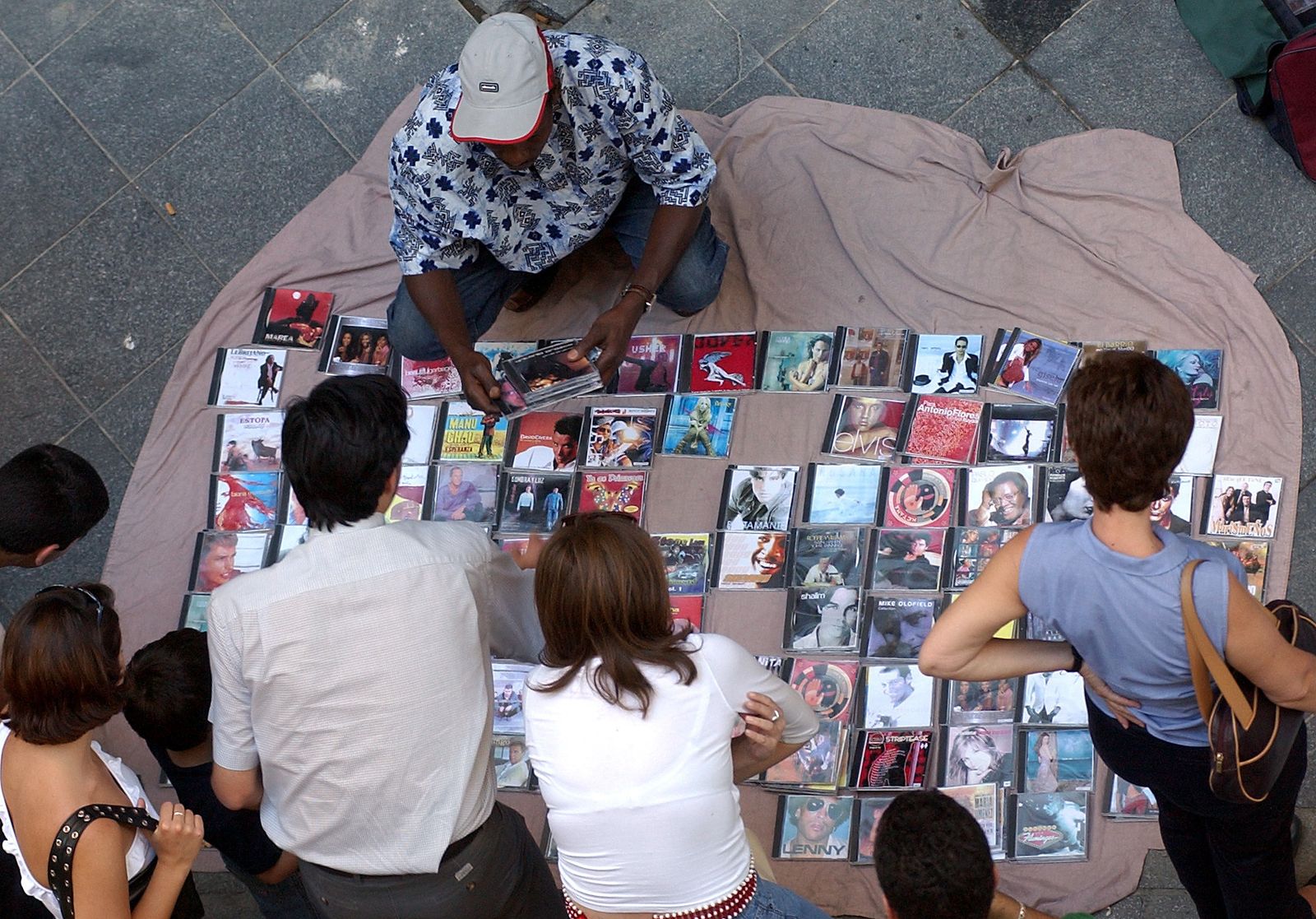 Un mantero ofrece copias de CDs a un grupo de personas en plena calle.