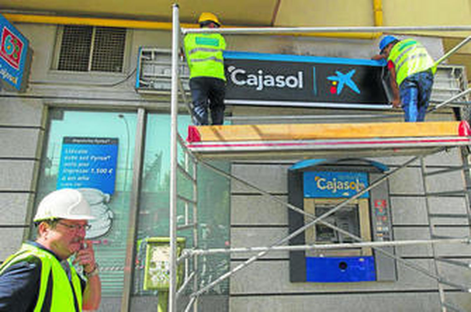 Operarios cambian el rótulo de Cajasol de una oficina por el que incluye el logo de La Caixa.