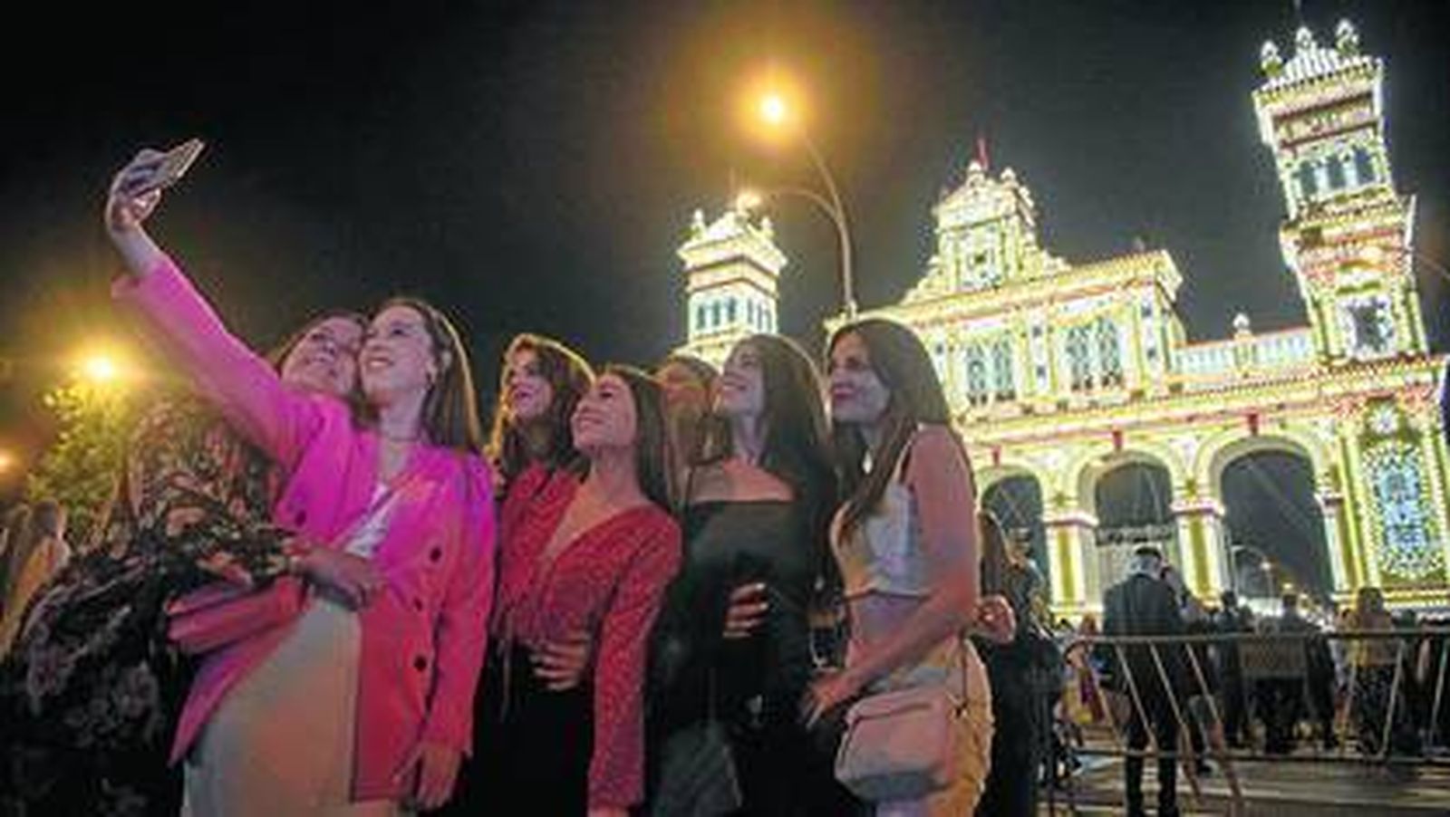 Varias jóvenes se hacen el primer selfi de la Feria con la portada al fondo tras el alumbrado.