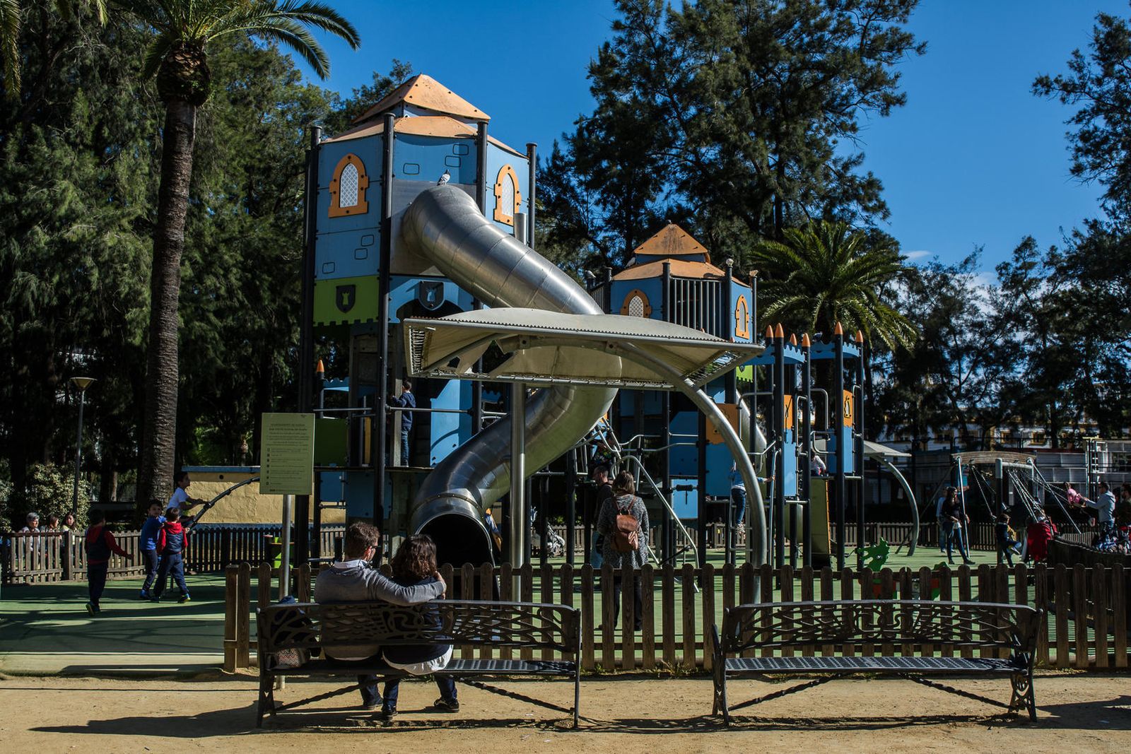 Una pareja contempla, sentada en un banco, como sus hijos disfrutan en uno de los parques infantiles que hay en la capital onubense.