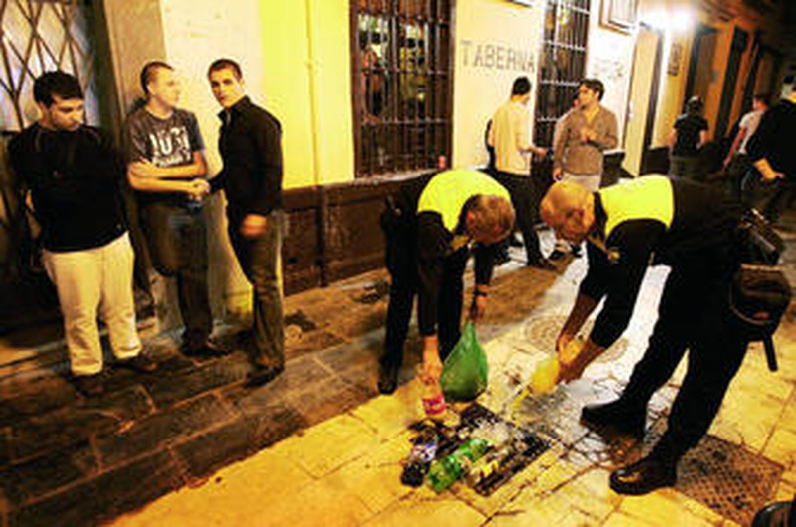 Dos policías locales tiran por una alcantarilla el alcohol de unos jóvenes concentrados en la Plaza Mitjana de la capital.