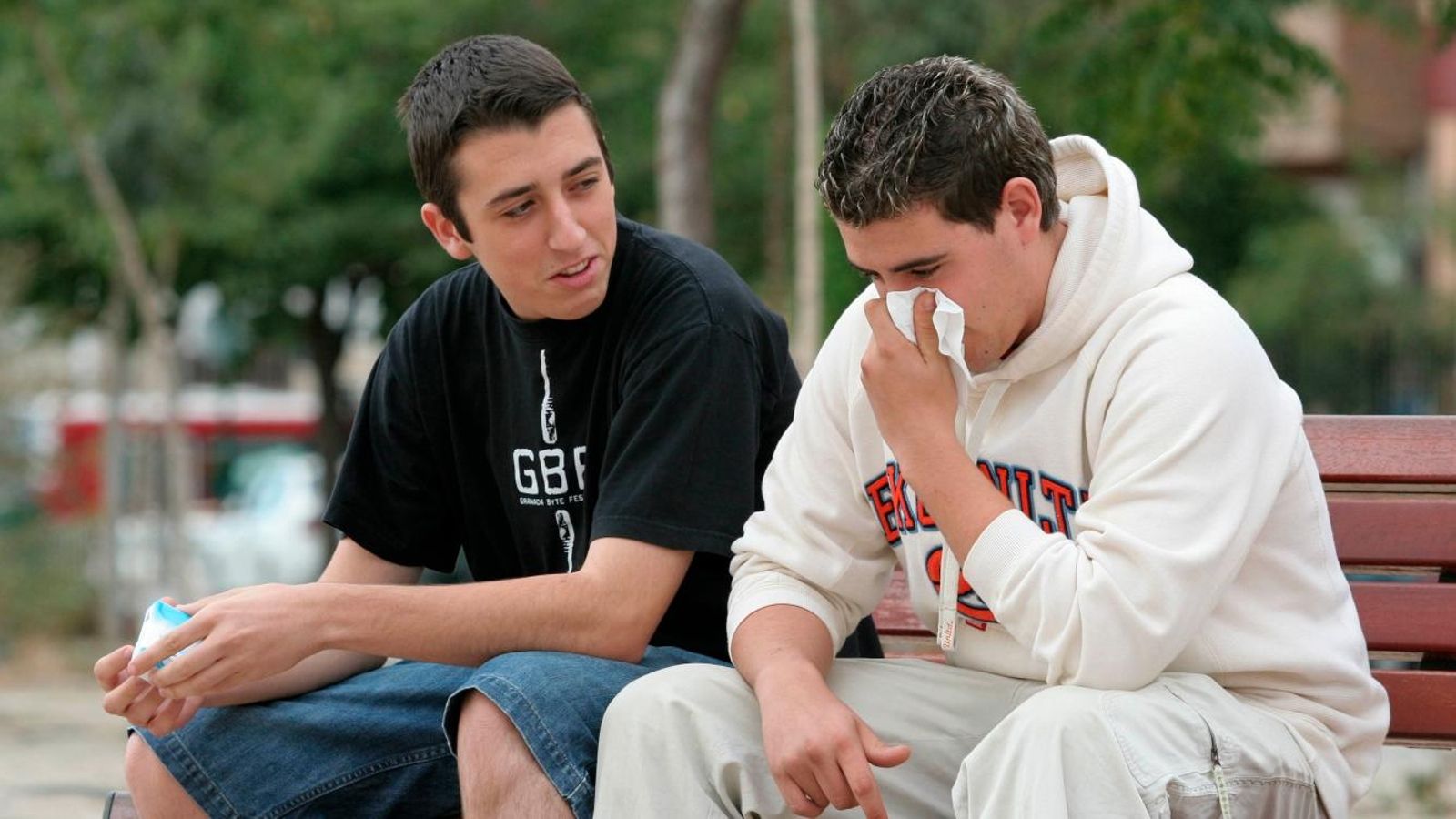 Dos jóvenes sentados en un banco, con uno de ellos sonándose los mocos.
