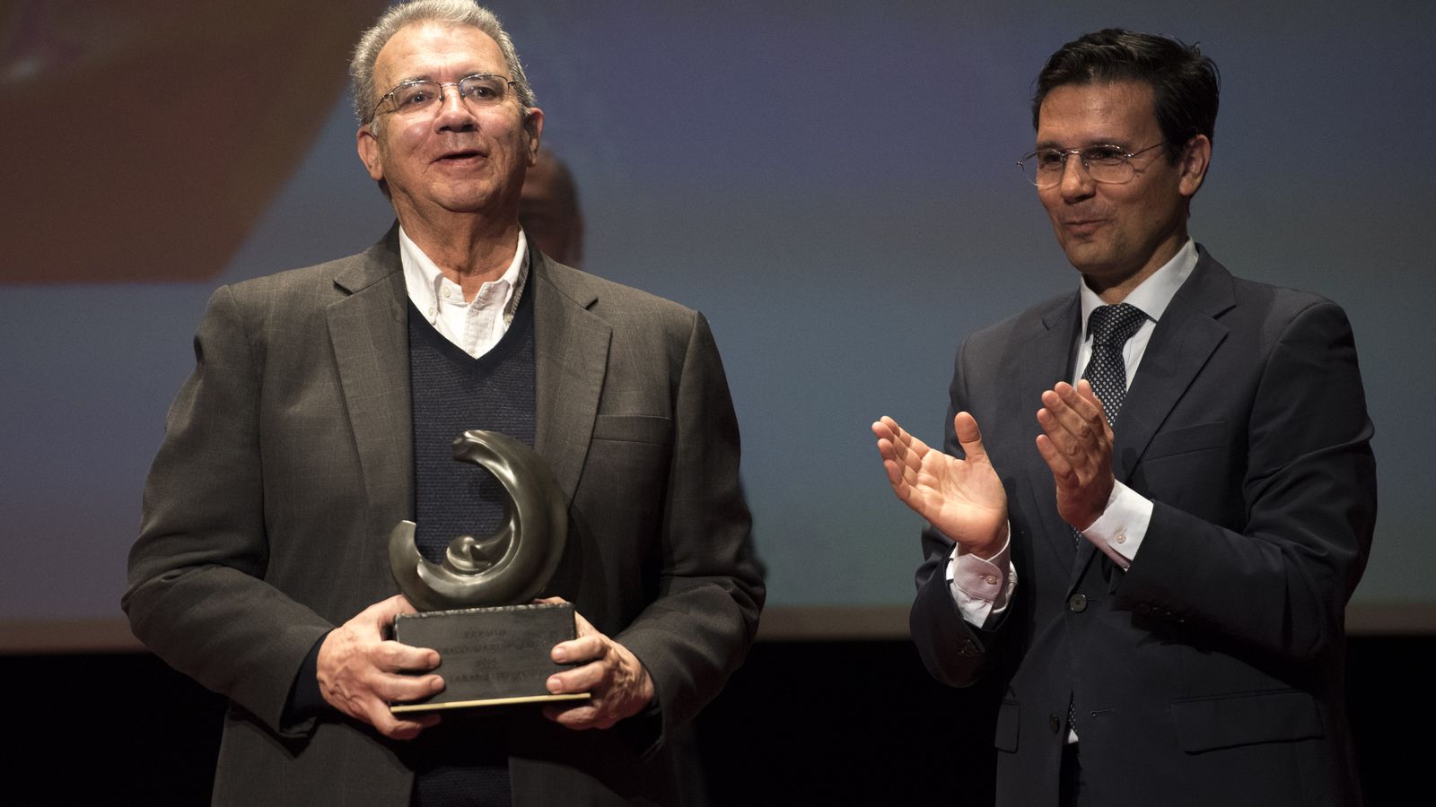 El escritor (a la izquierda) sostiene el trofeo del premio en forma de luna mientras Cuenca aplaude.