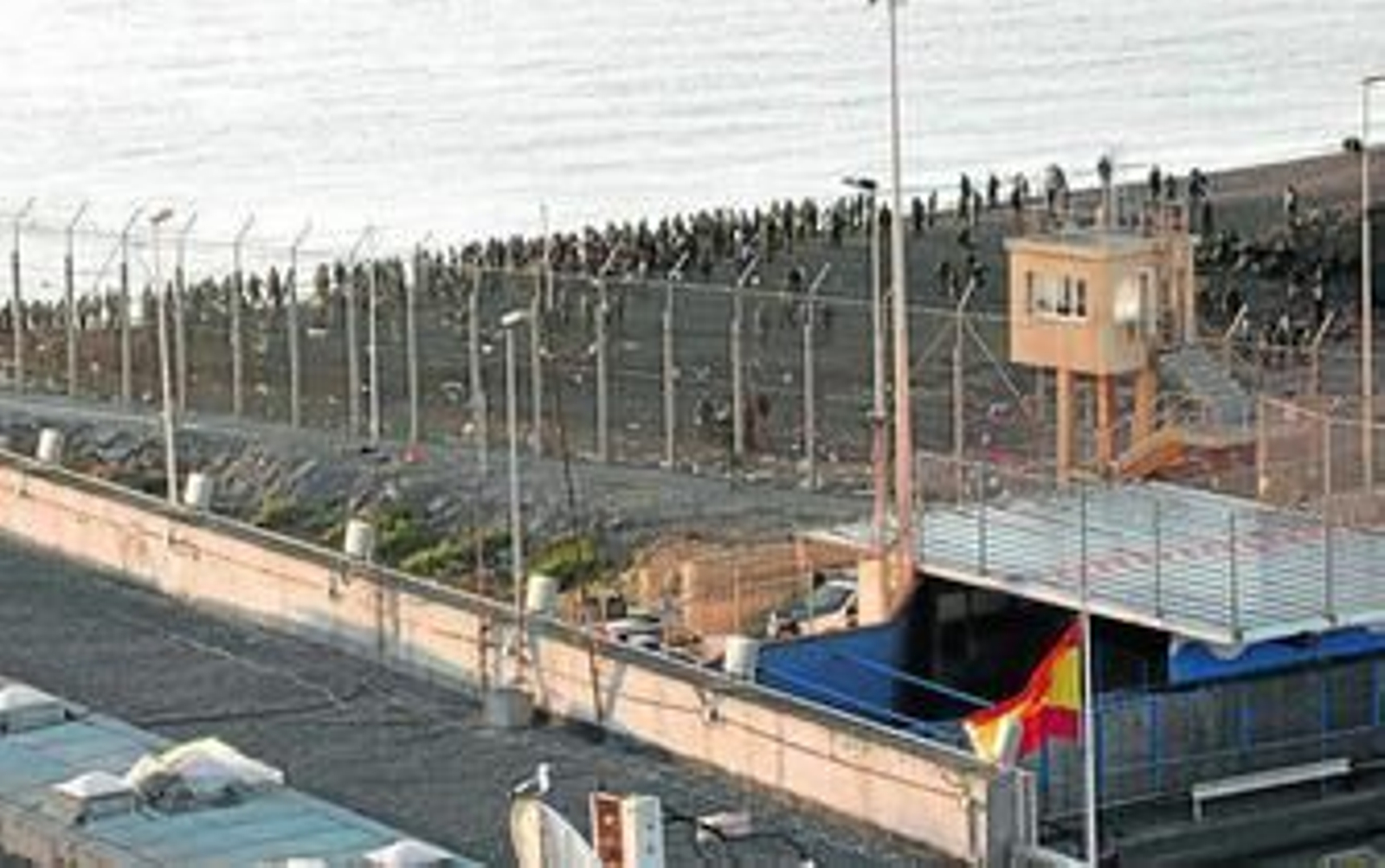 Valla de la frontera en Ceuta, con inmigrantes y policías marroquíes al otro lado.