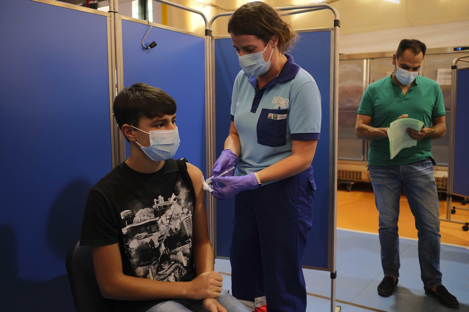 Un joven recibe la vacuna mientras su padre espera a pocos metros.