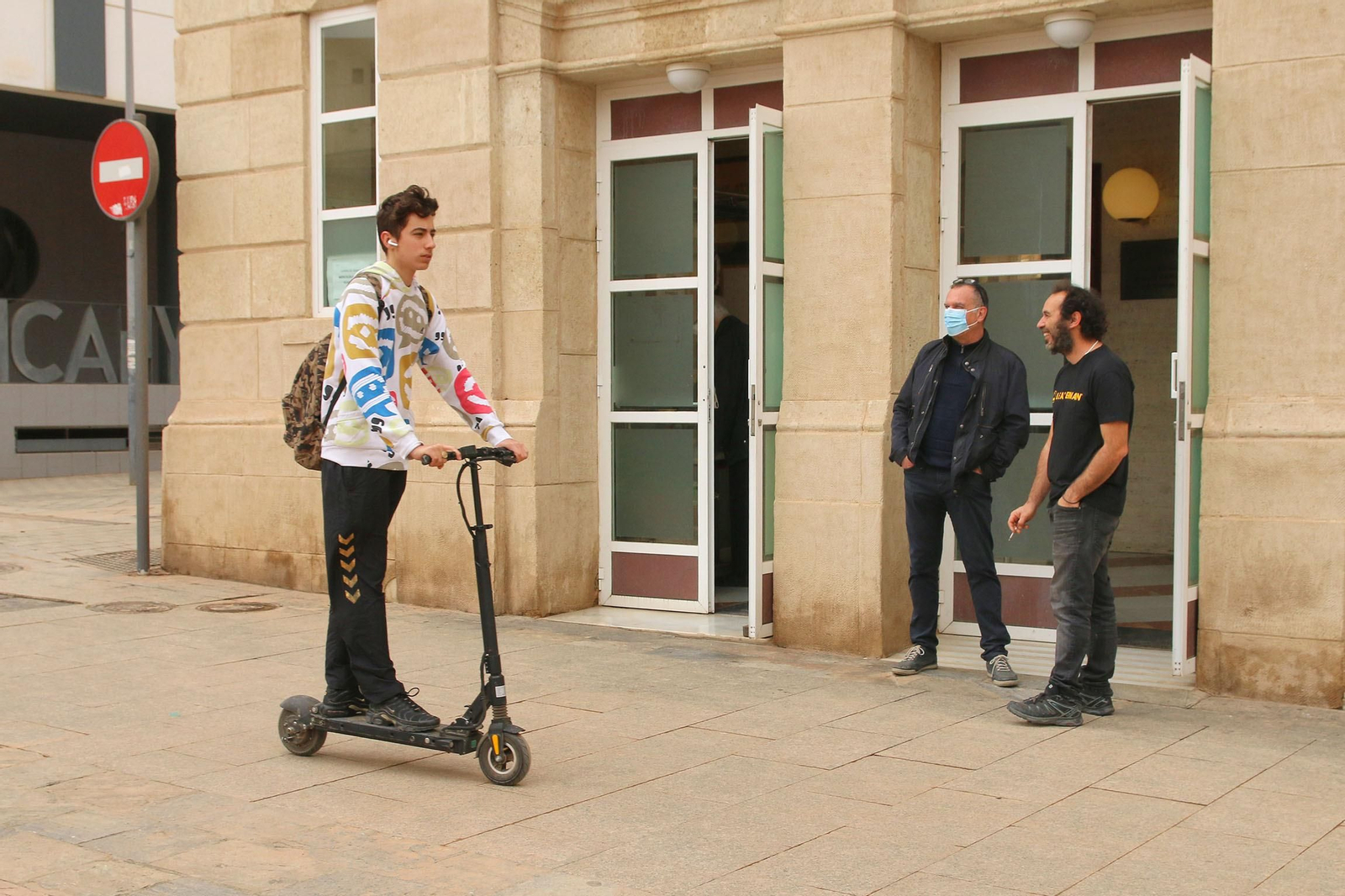 Joven en patinete, circulando por la acera, sin casco y con auriculares.