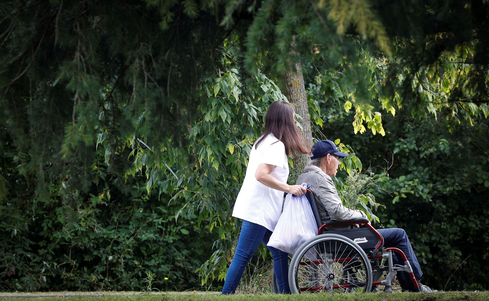 Una cuidadora pasea a un hombre en silla de ruedas por el jardín de una residencia. Una cuidadora pasea a un hombre en silla de ruedas por el jardín de una residencia.