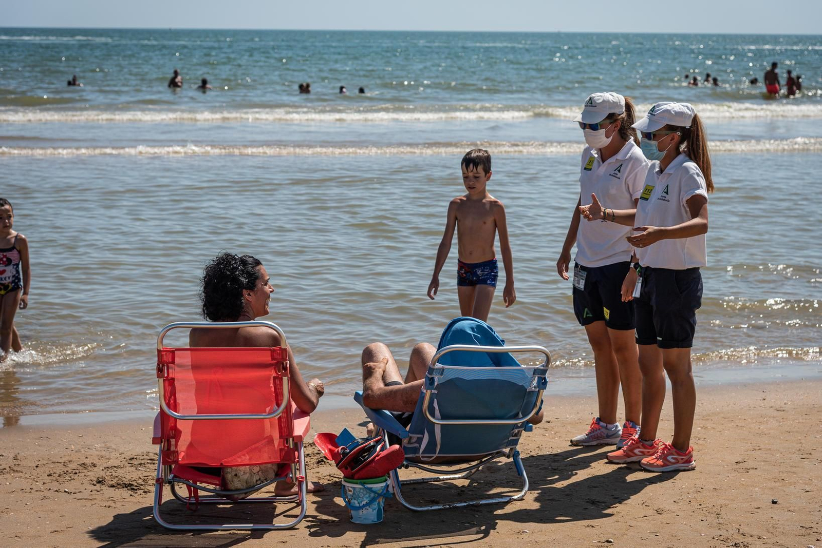 Unas auxiliares de playa informan a unos bañistas sobre el uso de la mascarilla.
