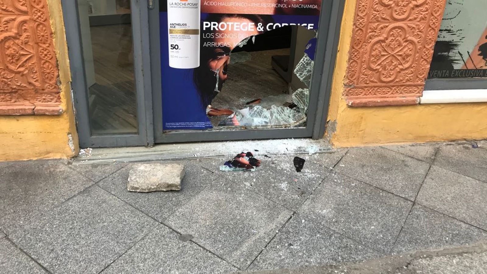 La puerta de la farmacia de Argote de Molina, tras el robo, y el adoquín con el que rompieron el cristal.