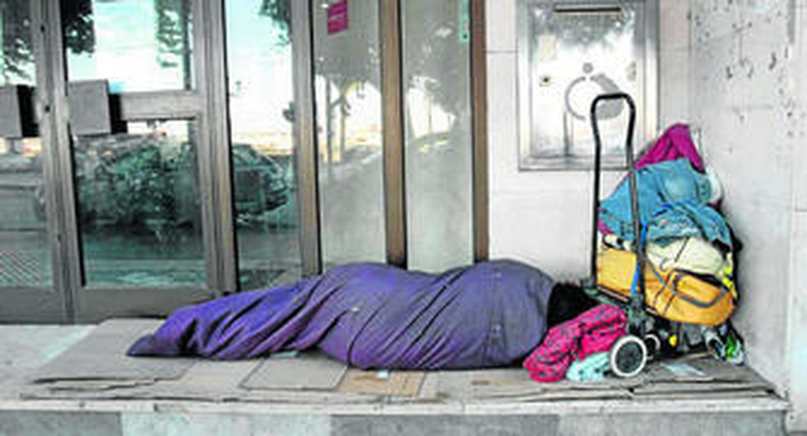 Una persona sin hogar duerme, junto a sus escasas pertenencias, en el escalón de entrada de un edificio de la ciudad.